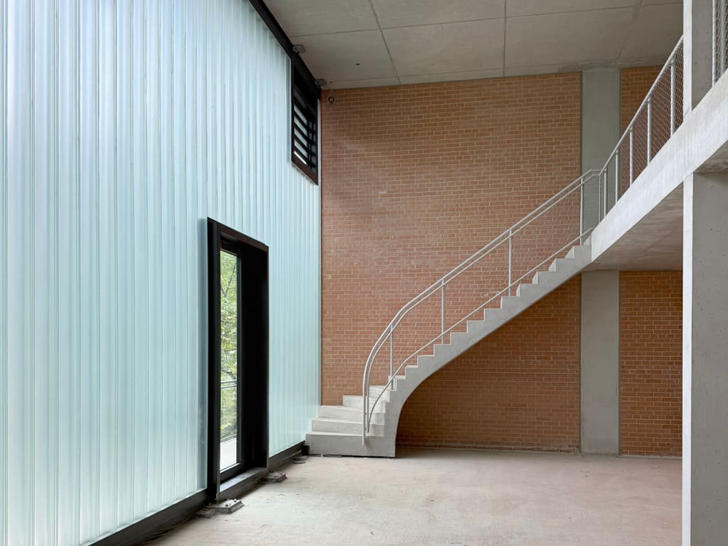 Rohbau der Halle mit geschwungener Betontreppe und weißem Rohrgeländer vor einer Klinkerwand, links die Wand aus Profilbauglas mit dunkler Glastür, rechts die Galerie mit Drahtgeflecht-Brüstung