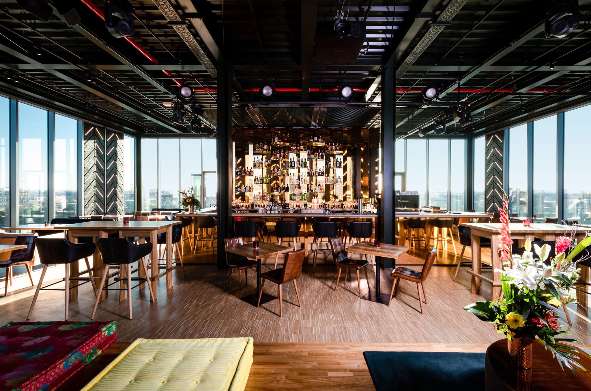 Rooftop-Bar mit Holztresen und beleuchteter Flaschenwand, umlaufende Panoramafenster über Berlin