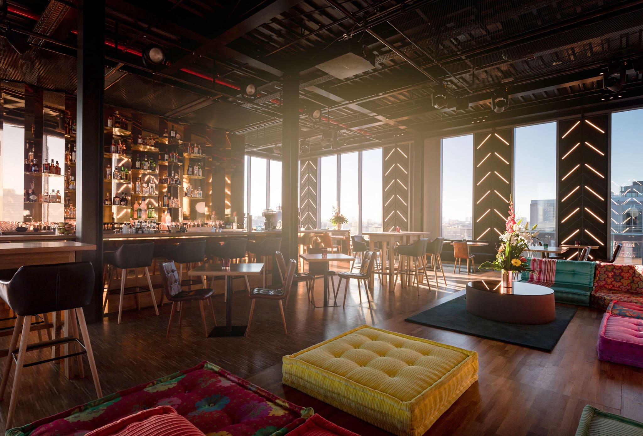 Bar im Gegenlicht der tiefstehenden Sonne, bunte Sitzkissen und gelber Sitzwürfel im Vordergrund