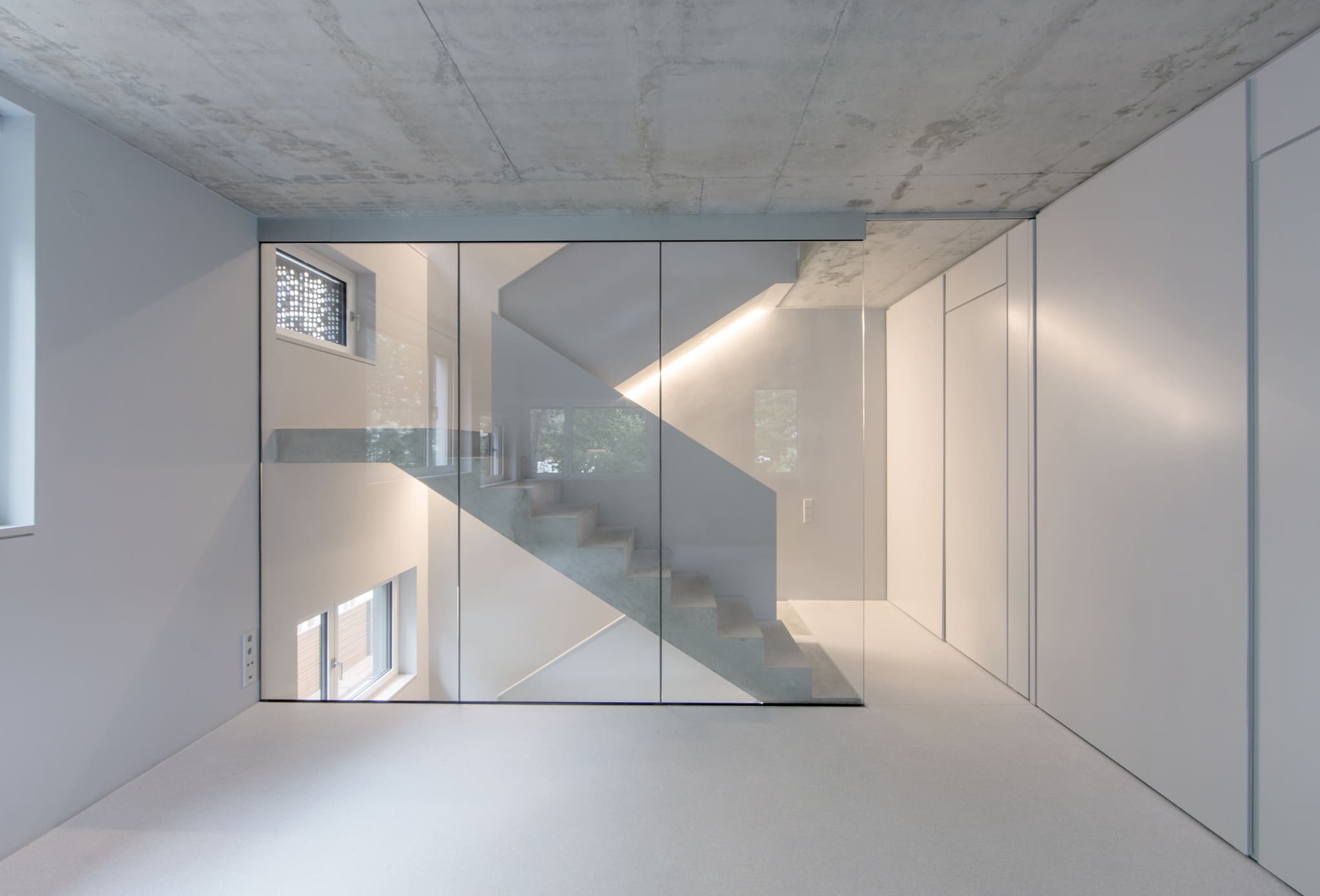 Raumhohe Glaswand vor einer Betontreppe, darüber Sichtbetondecke, rechts weiße Einbauschränke, heller Terrazzoboden