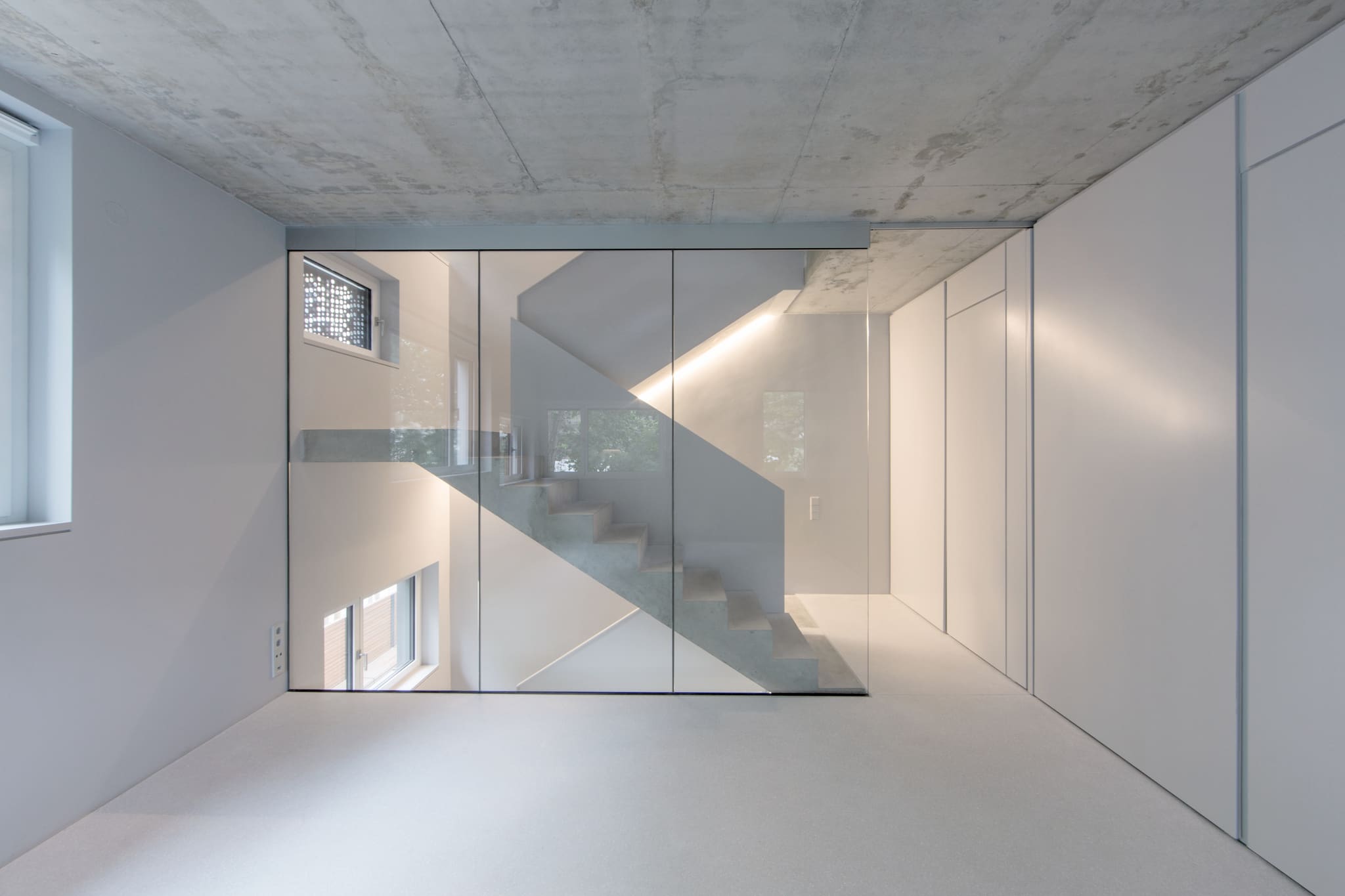 Full-height glass wall in front of a concrete stair, exposed concrete ceiling above, white fitted cupboards right, pale terrazzo floor
