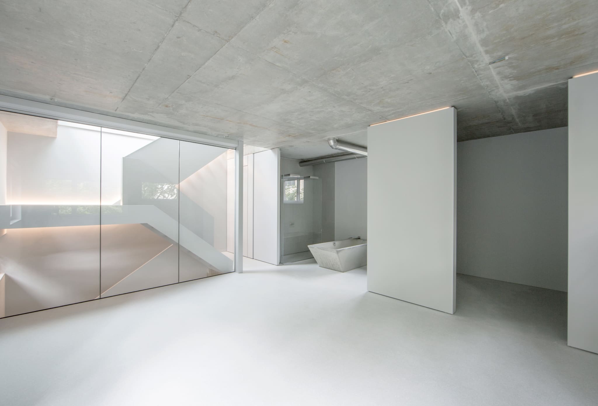 Offener Raum mit Sichtbetondecke, Glasabtrennung zum Treppenhaus, freistehender weißer Wandscheibe und Betonbadewanne im Hintergrund