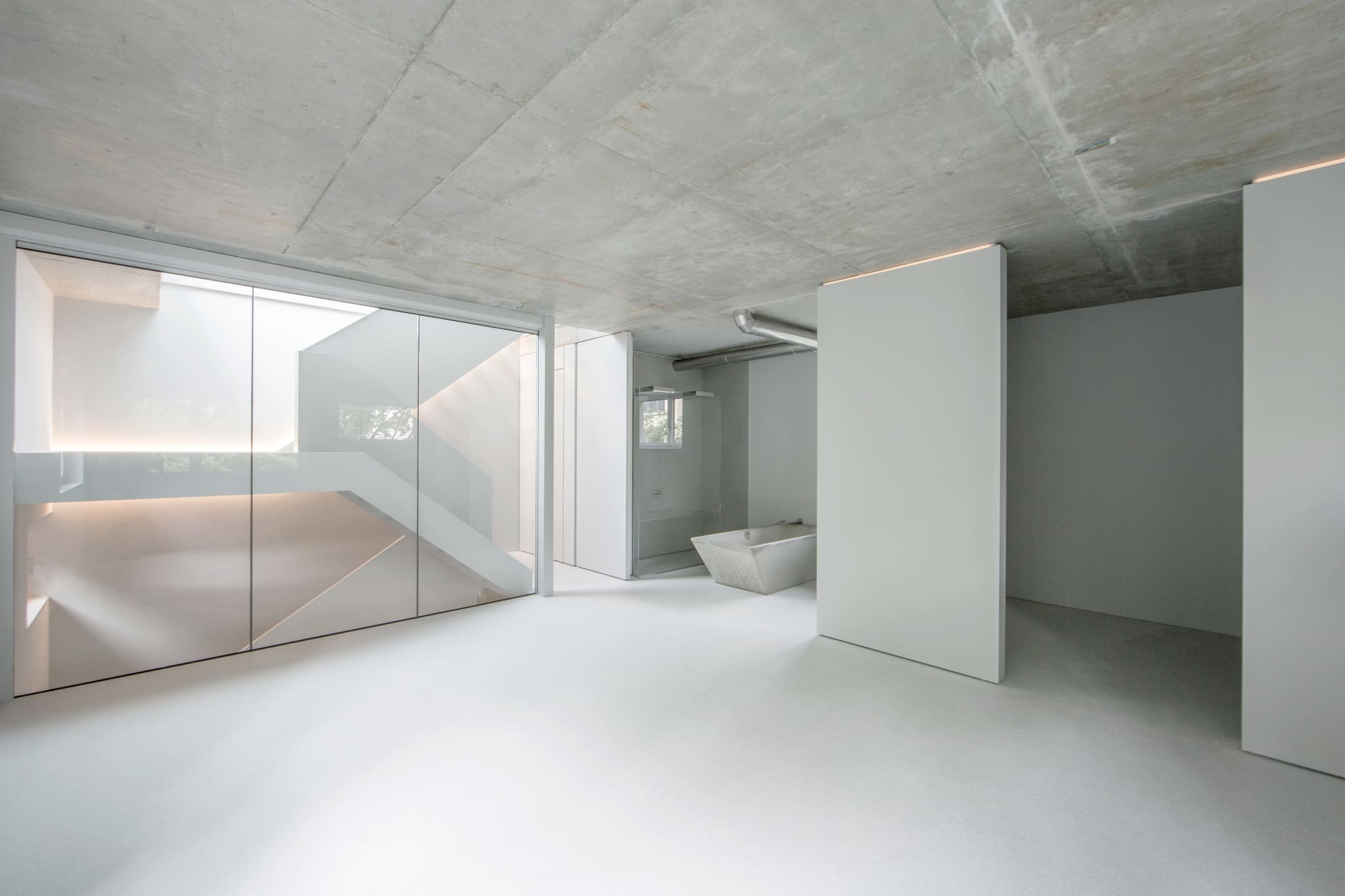 Open room with exposed concrete ceiling, glass enclosure to the stairwell, freestanding white wall panel and concrete bathtub behind