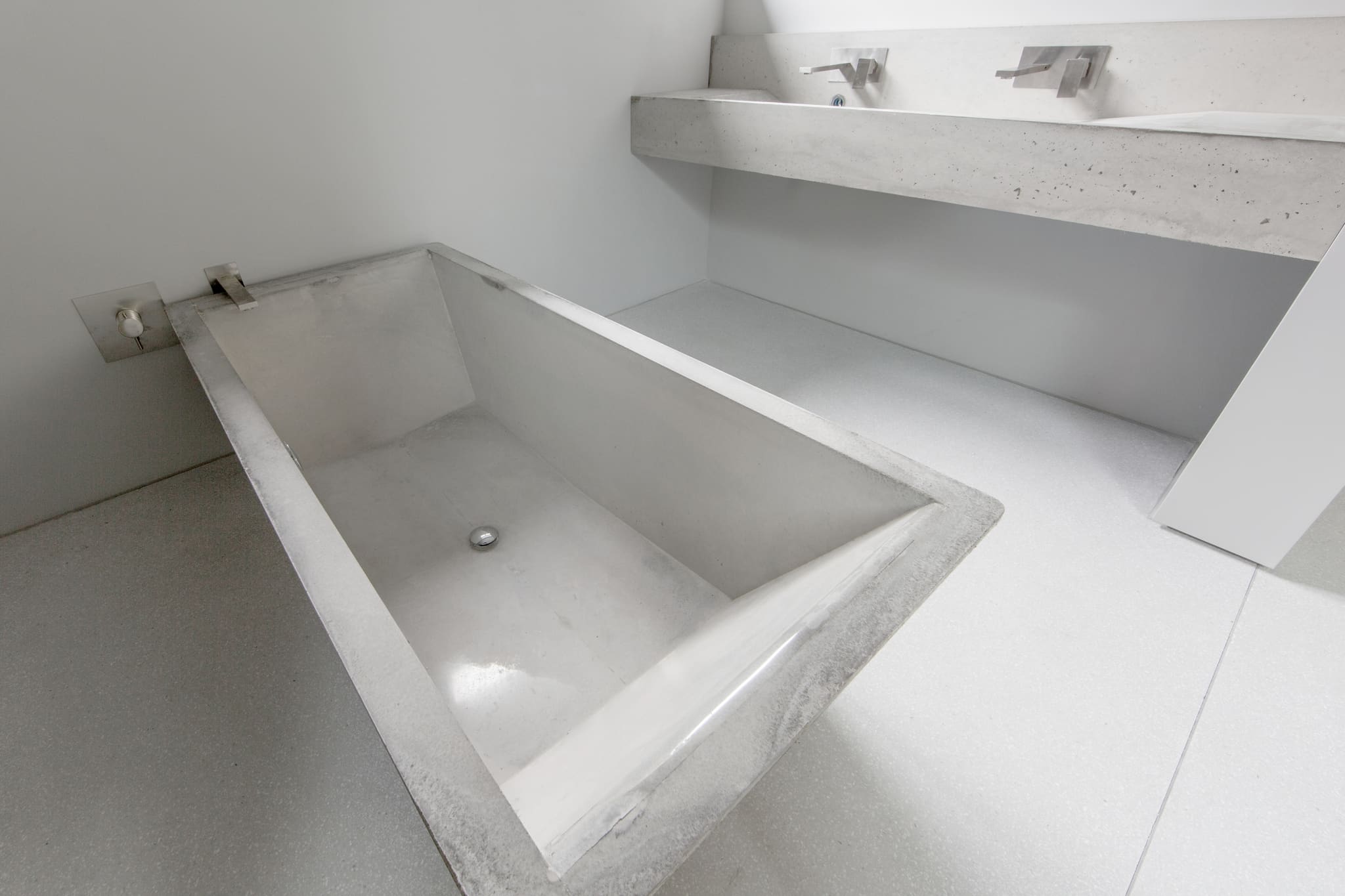 Rectangular concrete bathtub with sloping backrest, stainless steel wall spout, a concrete slab with basins above, terrazzo floor