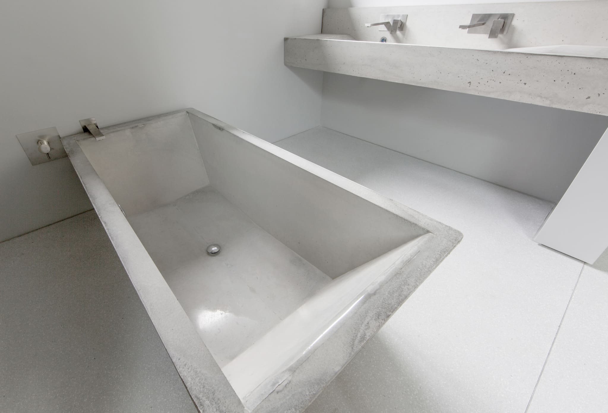 Rechteckige Betonbadewanne mit schräger Rückenlehne, Edelstahlarmatur an der Wand, darüber eine Betonplatte mit Waschbecken, Terrazzoboden