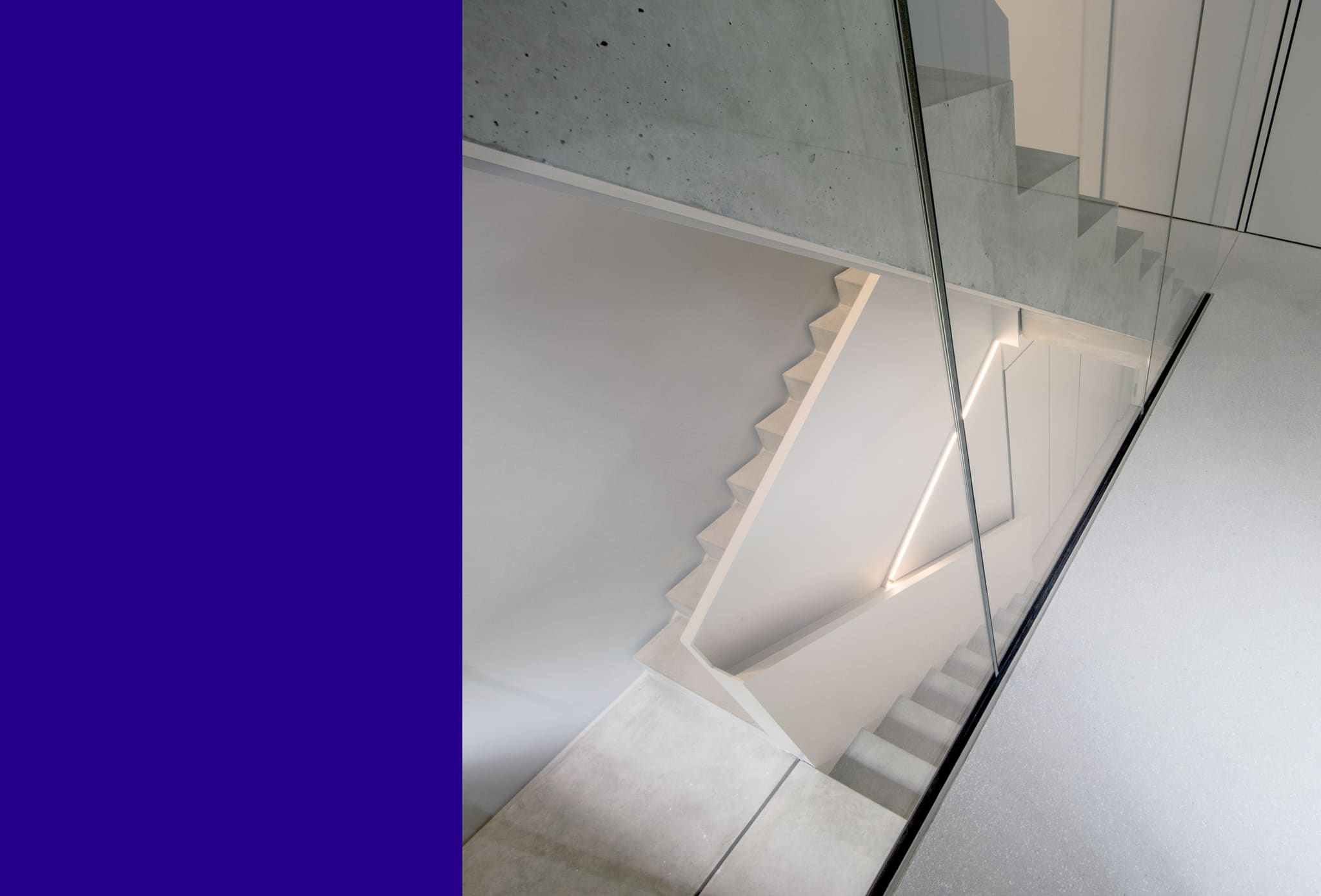 Blick von oben durch eine Glaswand auf eine Betontreppe mit beleuchteter Wange, Sichtbetondecke, links blaue Farbfläche