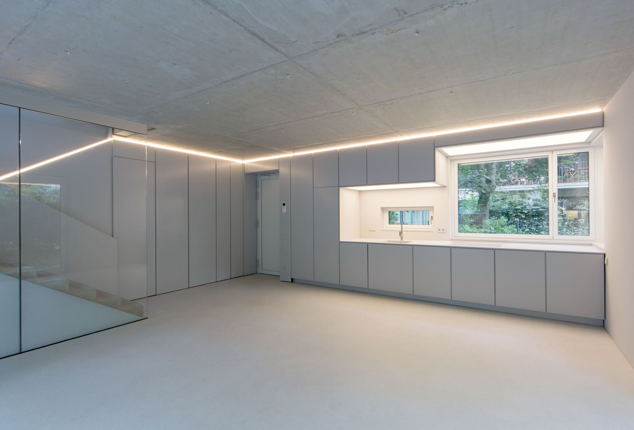 Raum mit hellgrauer Paneelwand, Küchenzeile unter dem Fenster, leuchtender Fuge unter der Sichtbetondecke und Glaswand zur Treppe