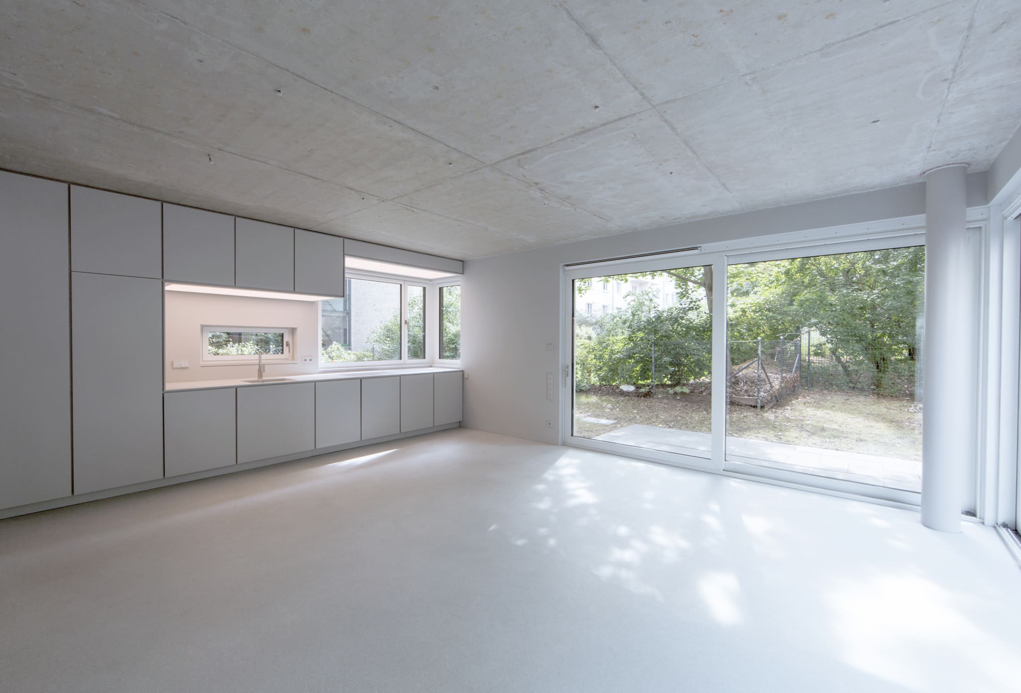 Leerer heller Raum mit Sichtbetondecke, grauer Schrankwand mit Küchenzeile, Terrazzoboden und raumhoher Schiebetür zum Garten