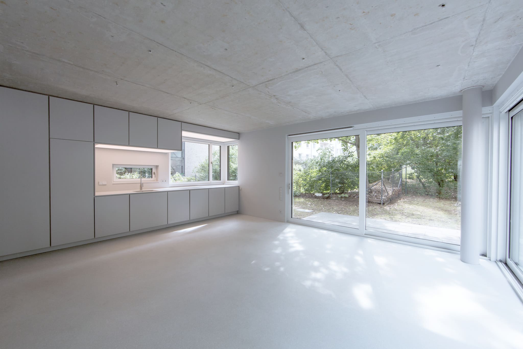Empty bright room with concrete ceiling, grey cupboard wall with kitchen run, terrazzo floor, full-height sliding door to the garden
