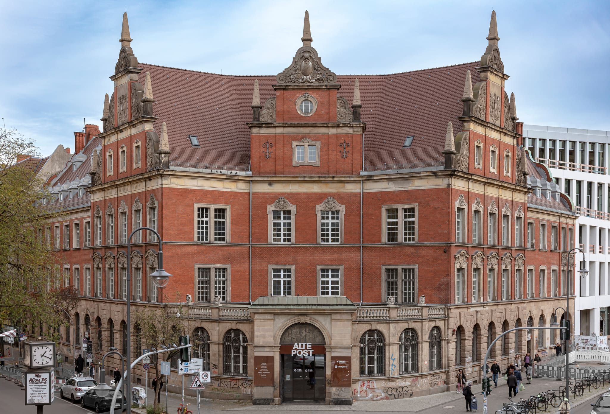 Historic brick building with scrolled gables, obelisks and mansard roof on a street corner, traffic lights and passers-by in front