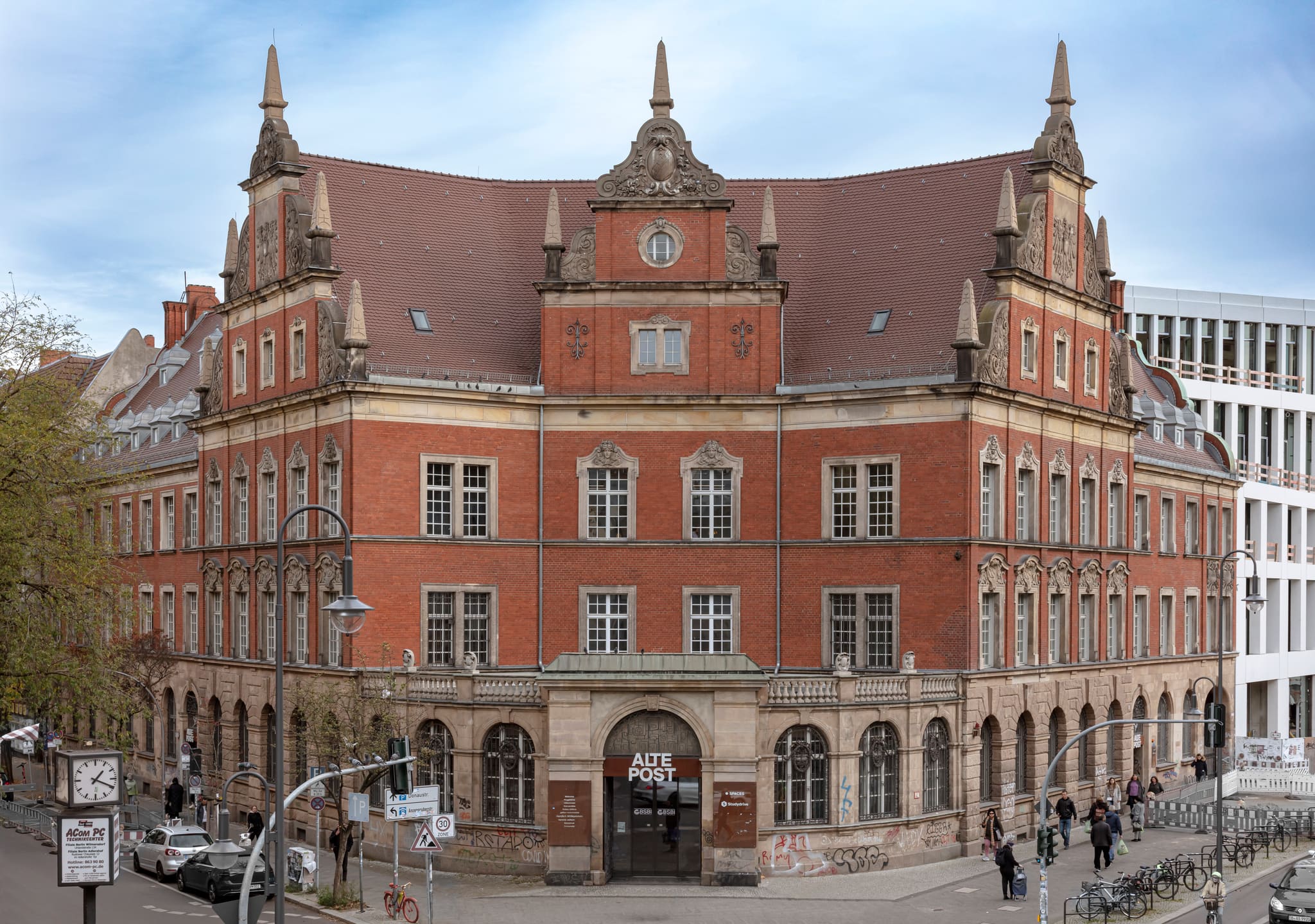 Historisches Backsteingebäude mit Schweifgiebeln, Obelisken und Mansarddach an einer Straßenecke, davor Ampeln, Radfahrer und Passanten