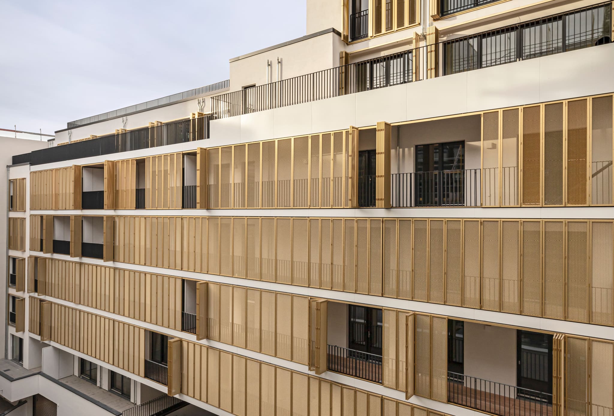 Oblique view of a pale facade with continuous balconies, gold perforated metal folding shutters in front, some panels folded open