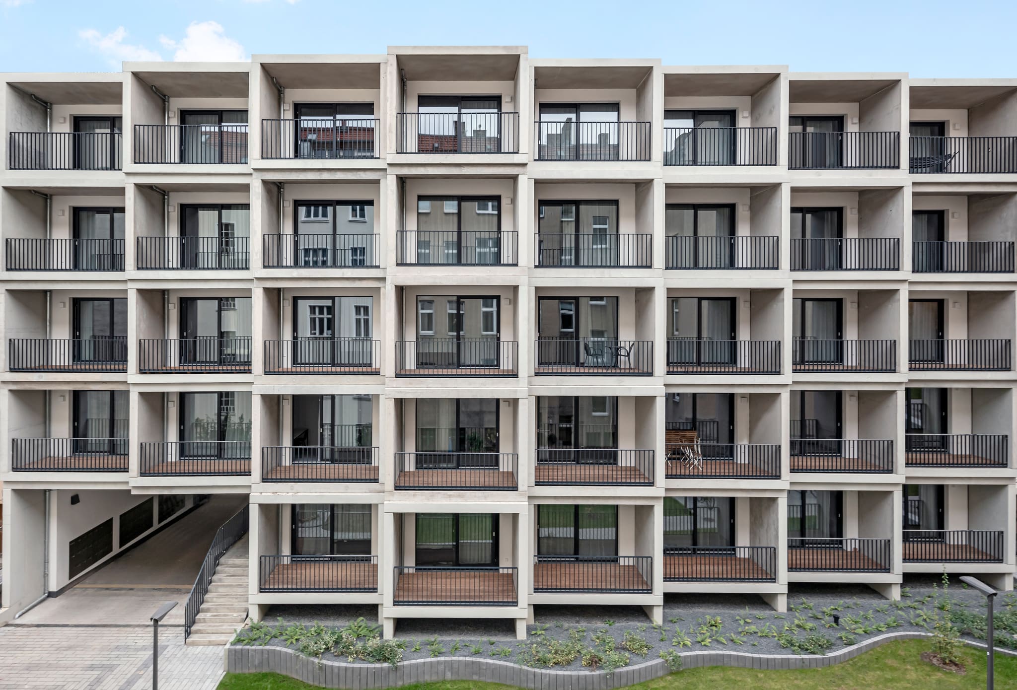 Five-storey facade of staggered exposed concrete boxes with timber decking, black bar railings and full-height windows, garage ramp below