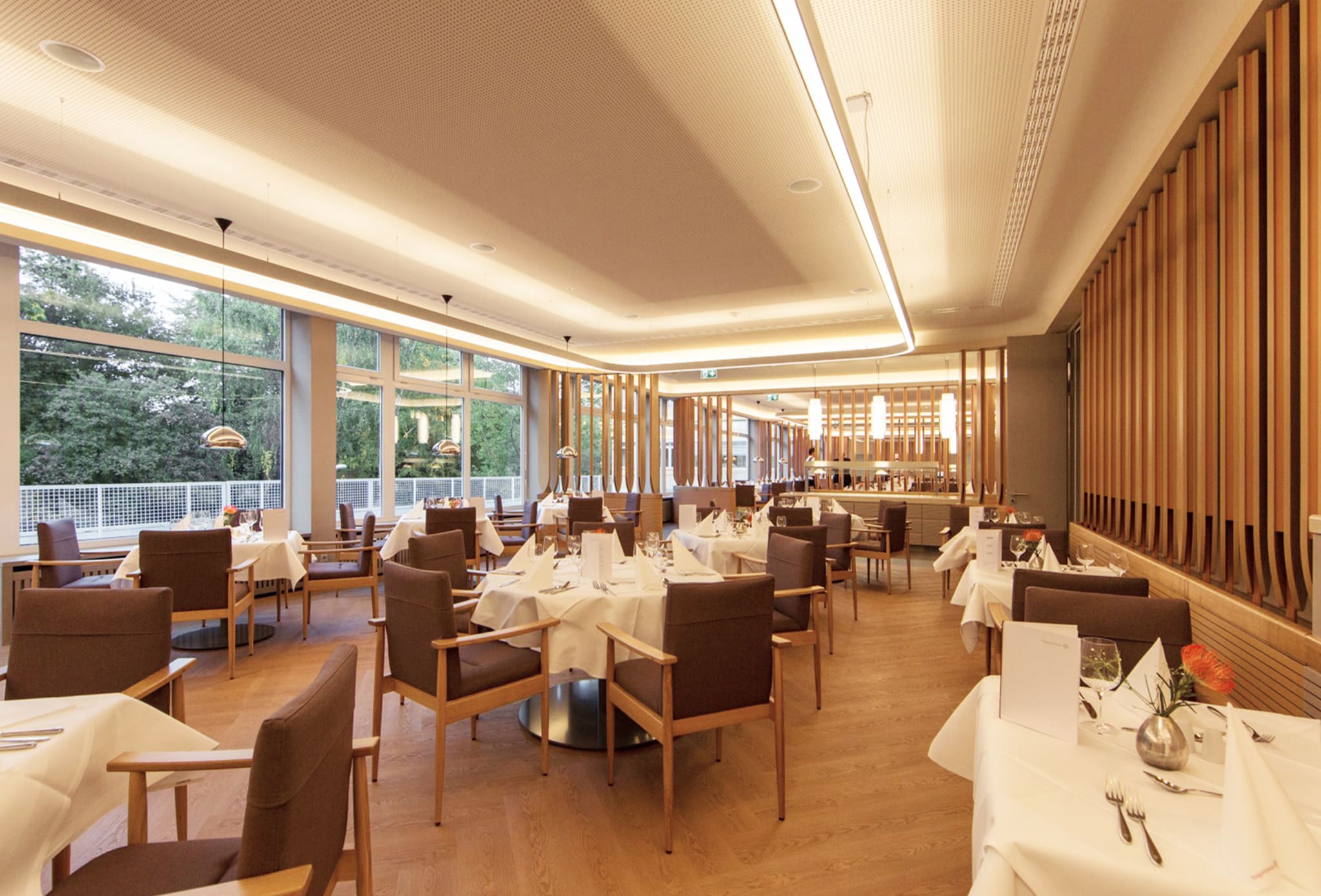 Restaurantsaal bei Tageslicht, raumhohe Fenster zum Grün, eingedeckte Tische mit Polstersesseln, rechts eine Wand aus Holzlamellen
