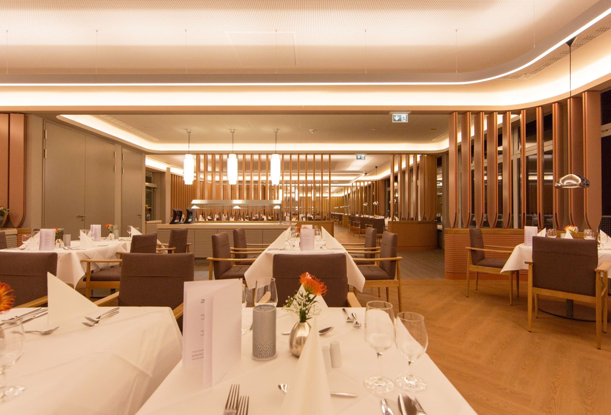 Blick über eingedeckte Tische auf eine zentrale Buffetinsel, darüber Pendelleuchten, seitlich Lamellenwände und umlaufende Lichtvoute