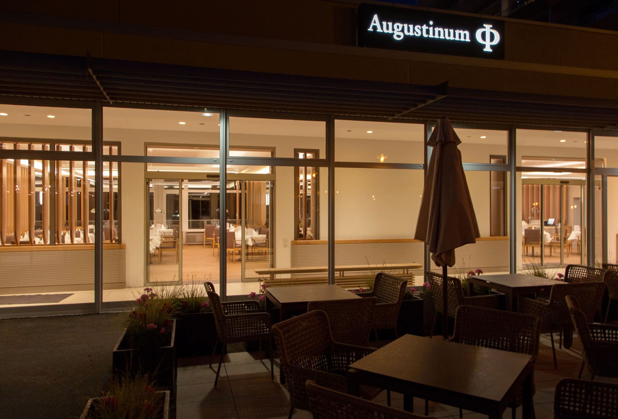Terrasse bei Nacht mit Korbsesseln, Tischen, geschlossenem Sonnenschirm und Pflanzkübeln vor der beleuchteten Glasfassade des Restaurants