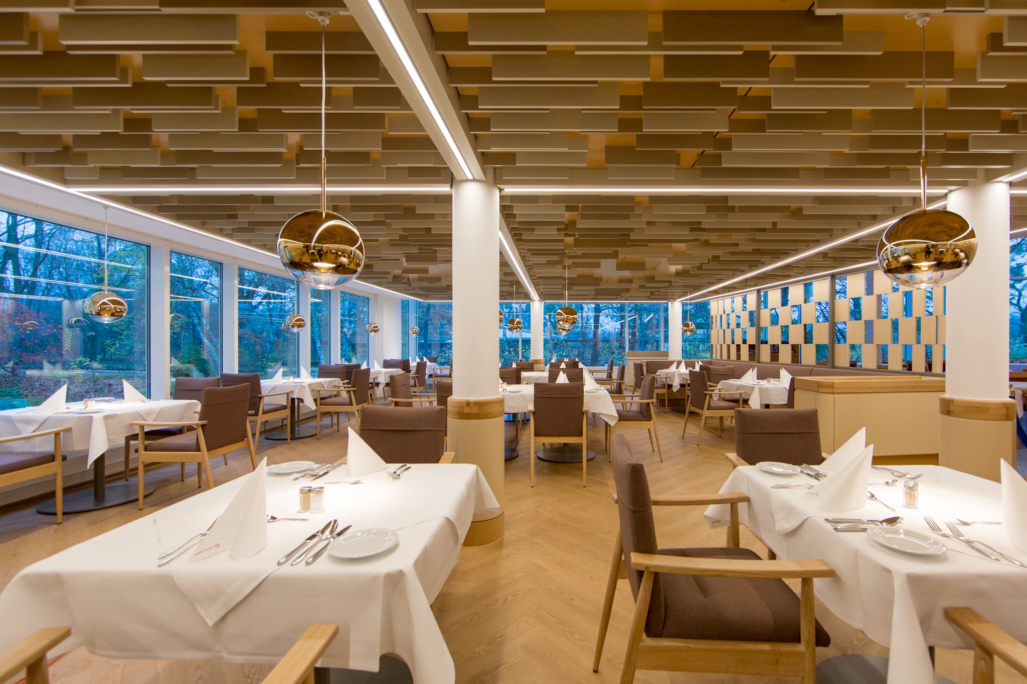 Gastraum mit runden Stützen, Messing-Kugelleuchten und Fensterfront ins Grüne unter versetzten Holzlamellen