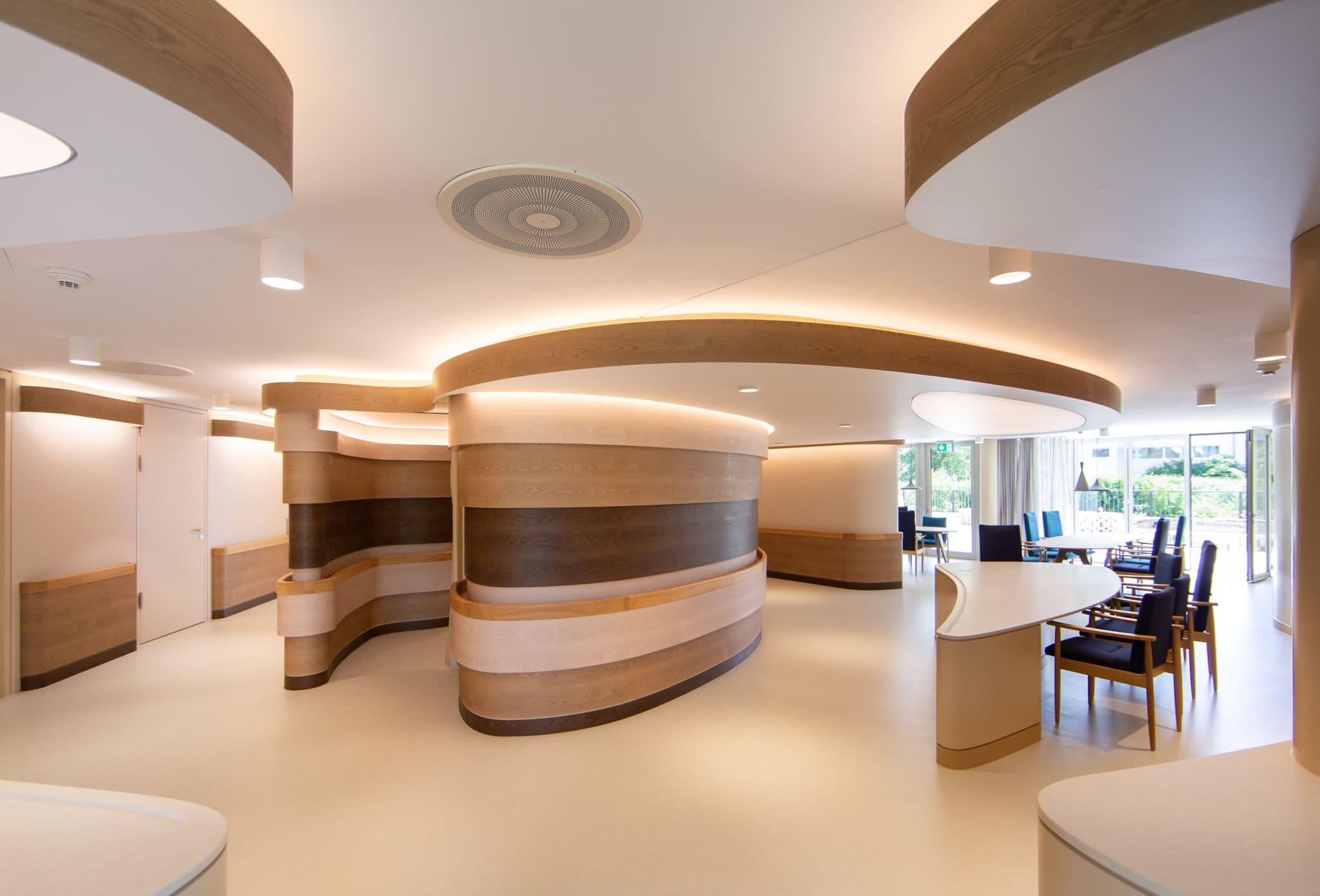 Open lounge area with wave-shaped timber walls and round ceiling discs, dining places with blue chairs at full-height glazing