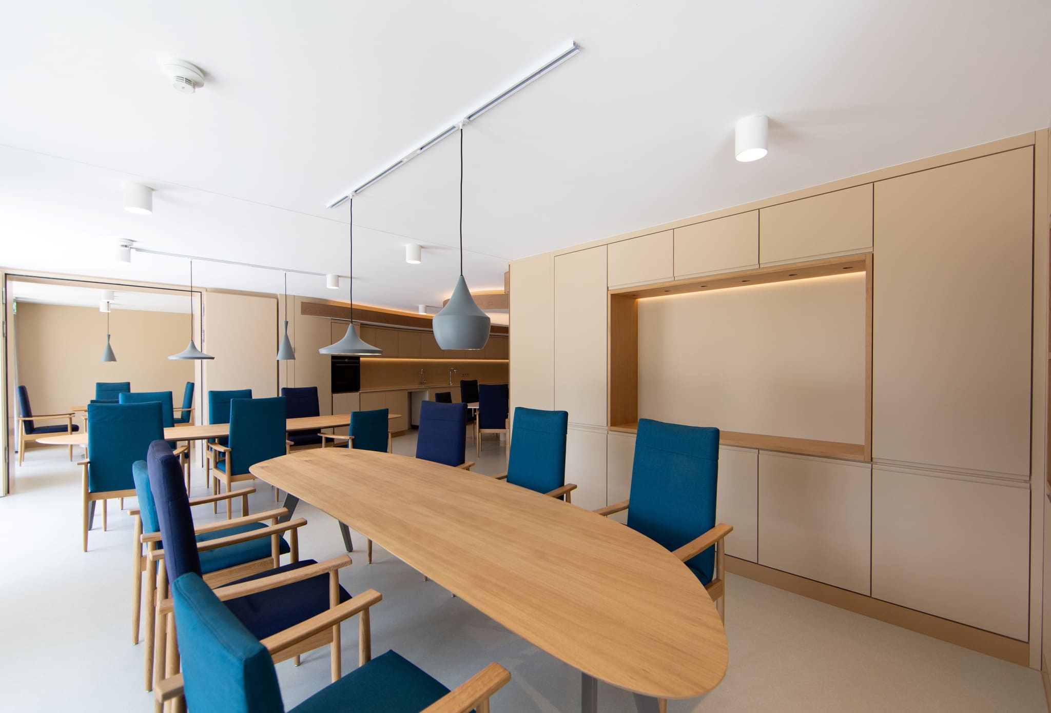 Dining area with long oval oak table, blue armchairs, sand-coloured cupboard wall with lit niche, open kitchen behind
