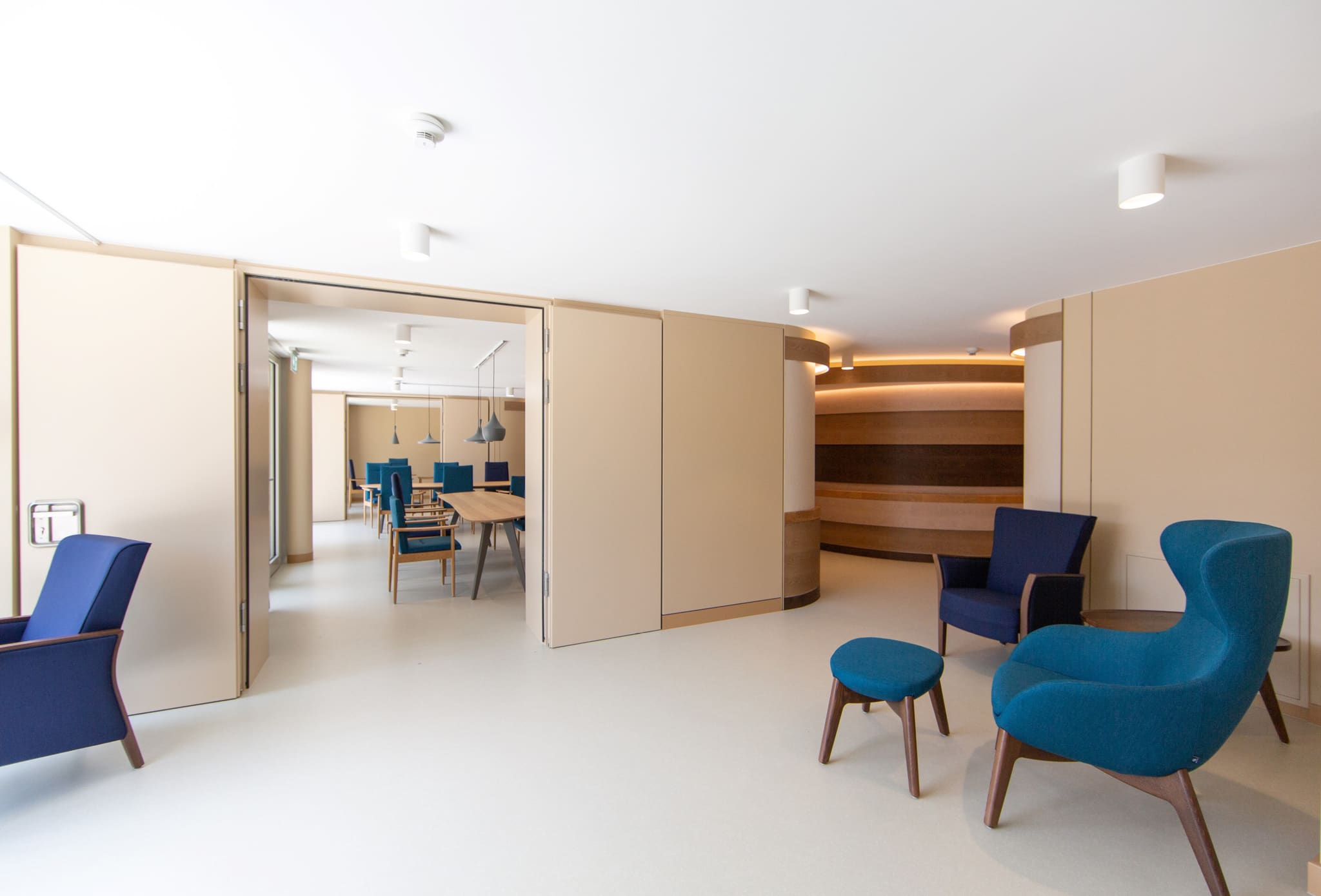 Seating area with blue armchairs and stool, opened folding wall revealing dining tables, wave-shaped timber wall at right