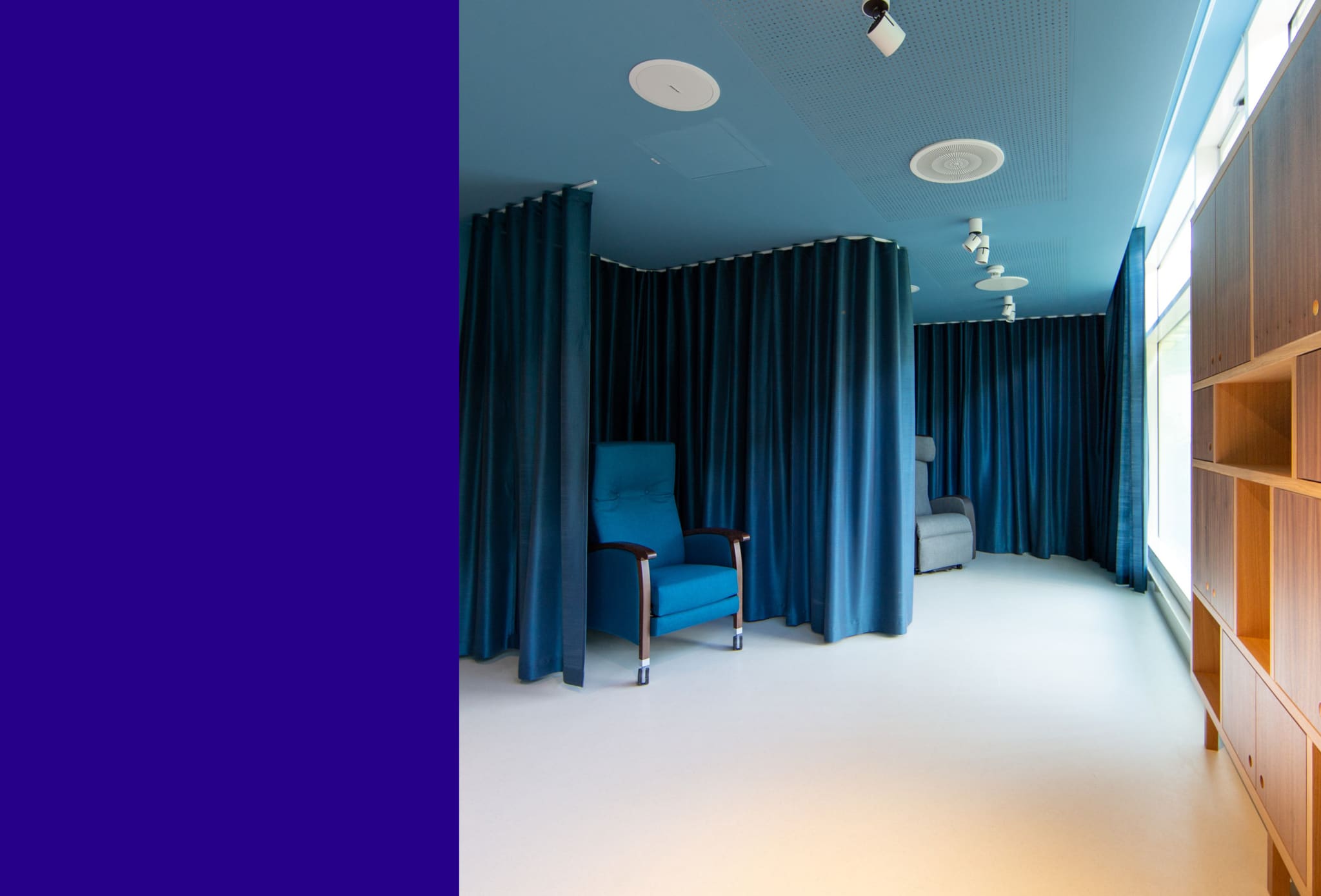 Rest room with blue ceiling, teal curtains forming alcoves around two reclining chairs, oak shelving at a bright window wall