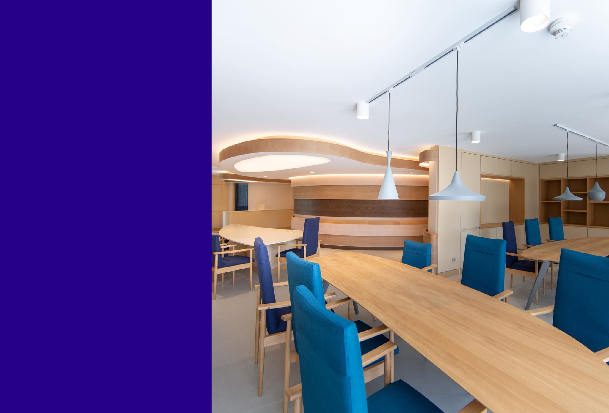 Dining area with long oak tables and blue chairs, pendant lamps, wave-shaped timber wall and lit ceiling sail behind