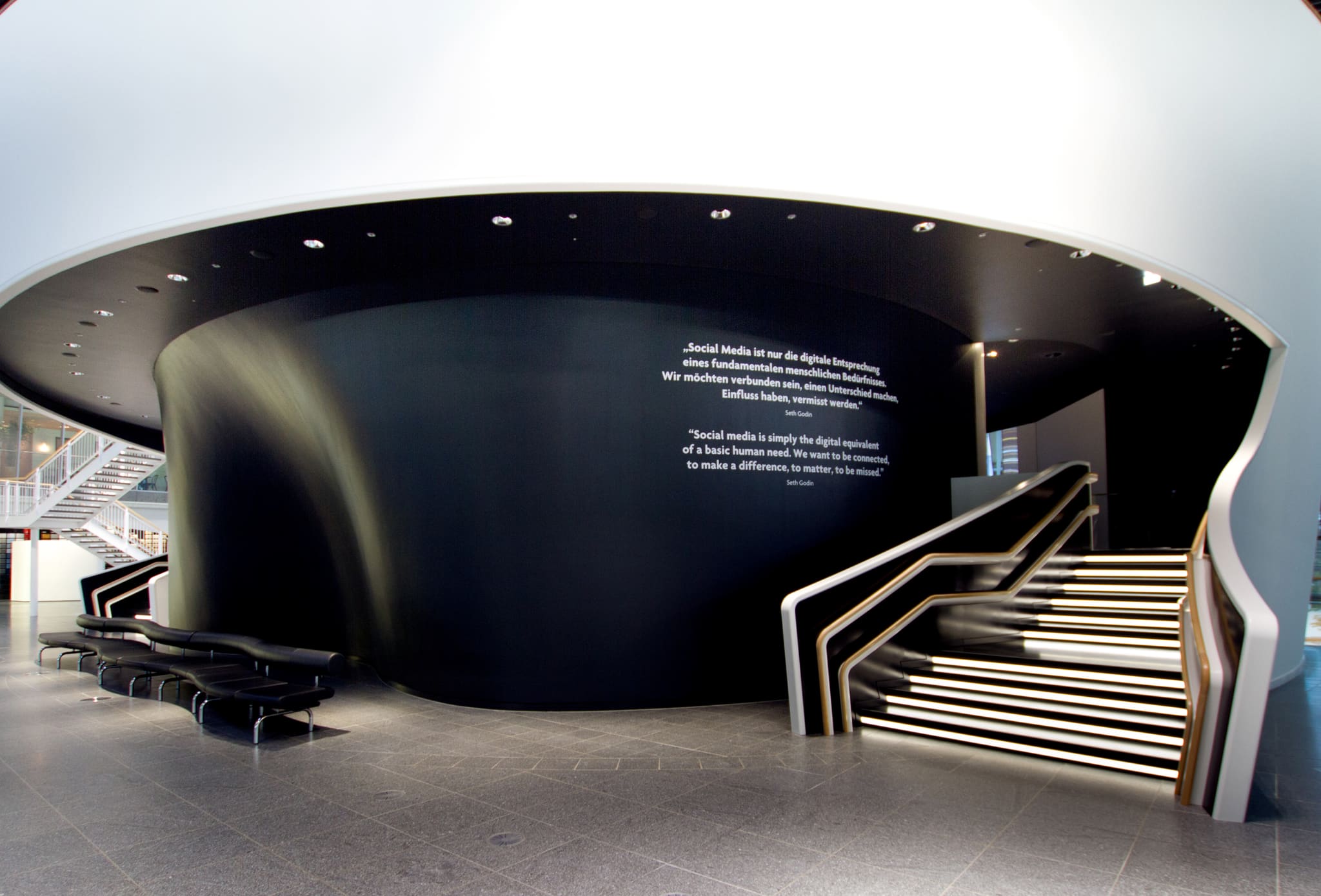 Foyer mit schwarzer, doppelt geschwungener Wandschale unter weißer Deckenscheibe, rechts Treppe mit weißer Brüstung und beleuchteten Stufen, links Sitzbänke