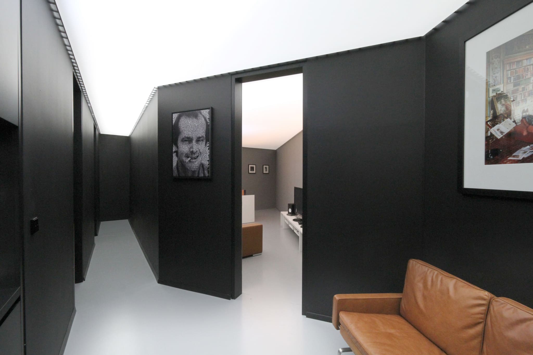 Corridor with black angled wall slabs beneath a glowing white ceiling, pale grey floor, framed portrait, brown leather sofa