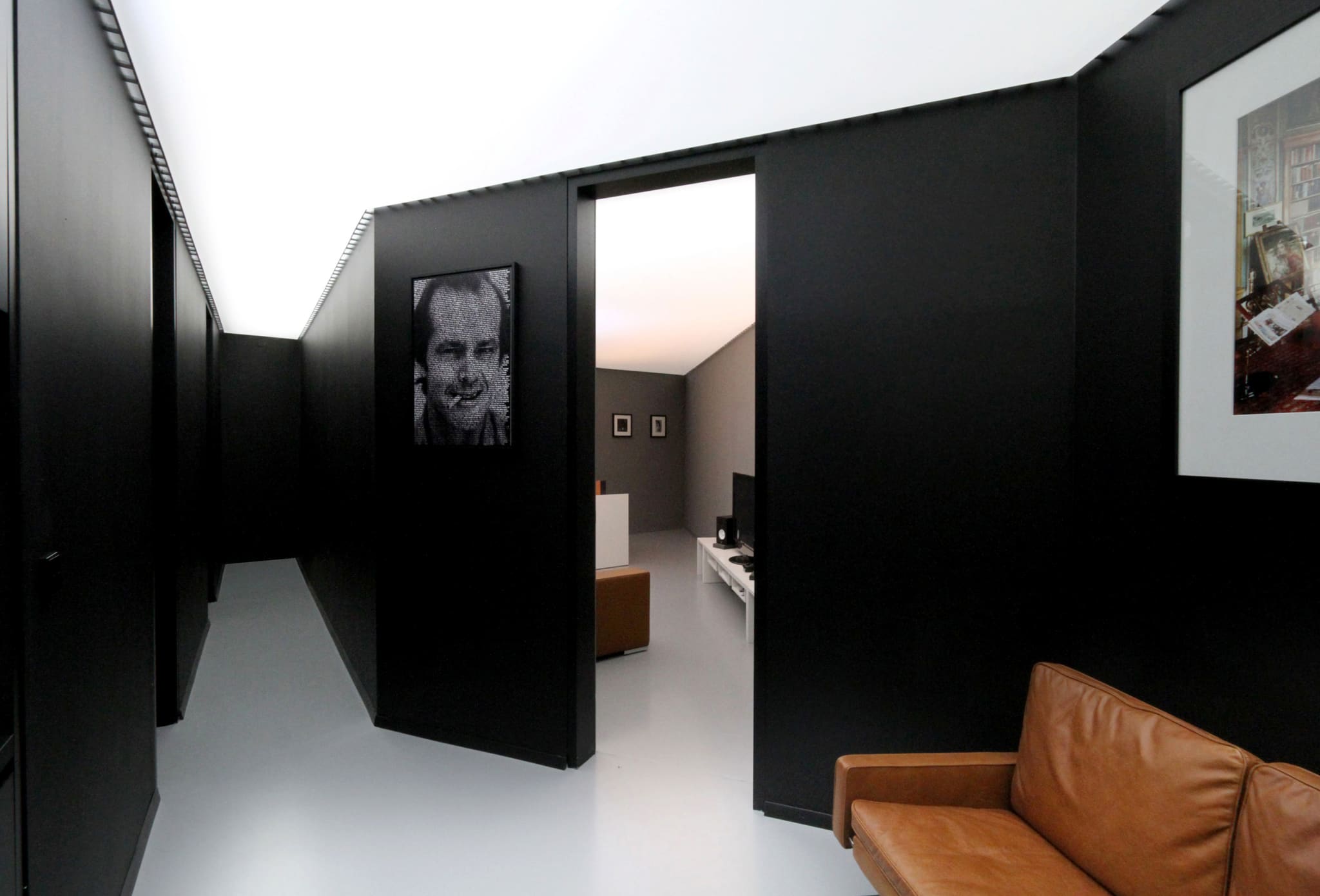 Corridor with black angled wall slabs beneath a glowing white ceiling, pale grey floor, framed portrait, brown leather sofa