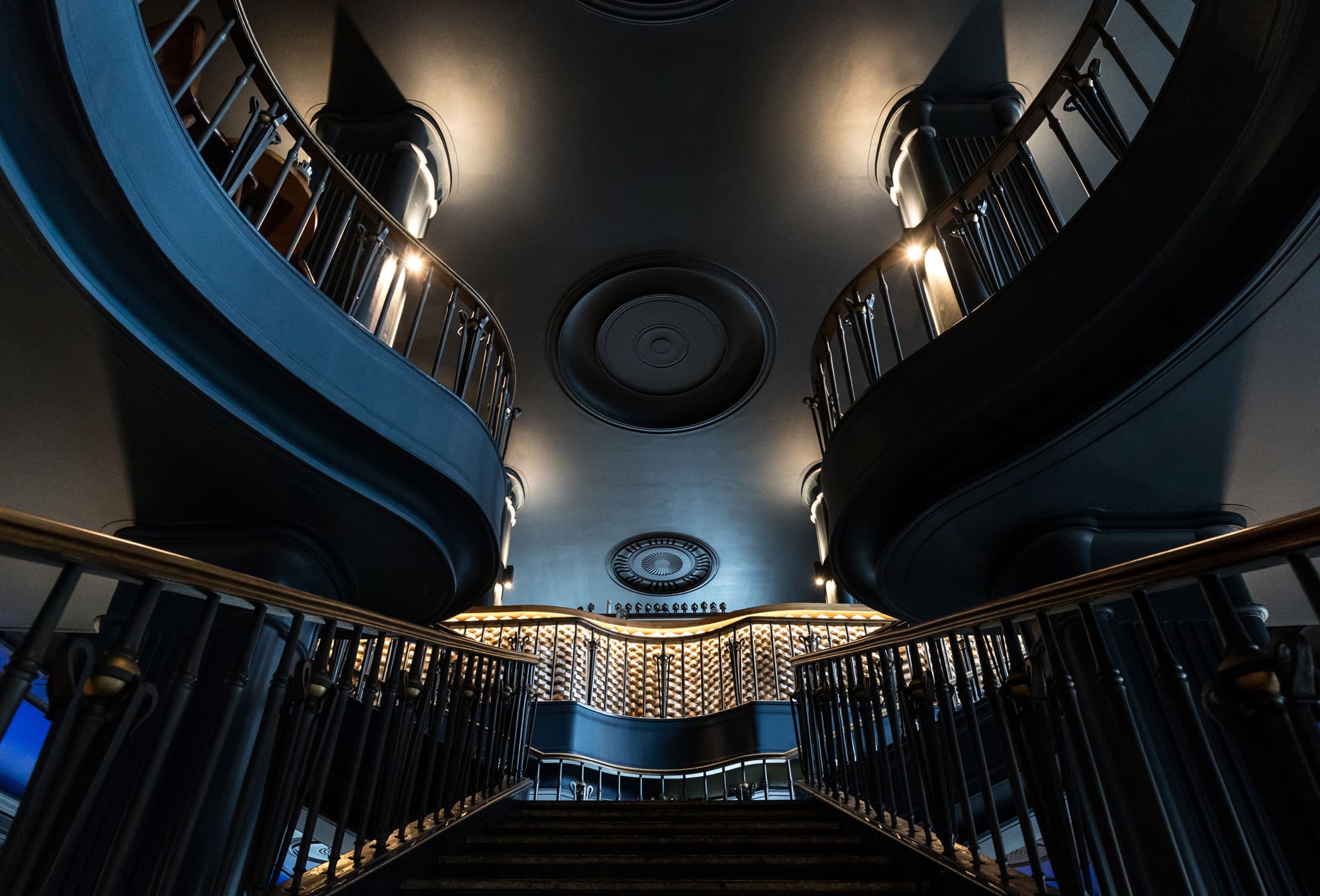 View up the curved open staircase, dark blue walls and ceiling rosettes, backlit brick counter behind a wrought iron balustrade