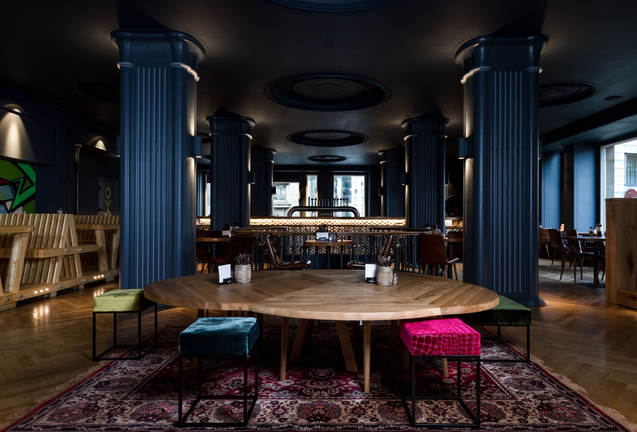 Dining room with dark blue columns, round timber table on an oriental rug, colourful velvet stools, lit brick counter behind