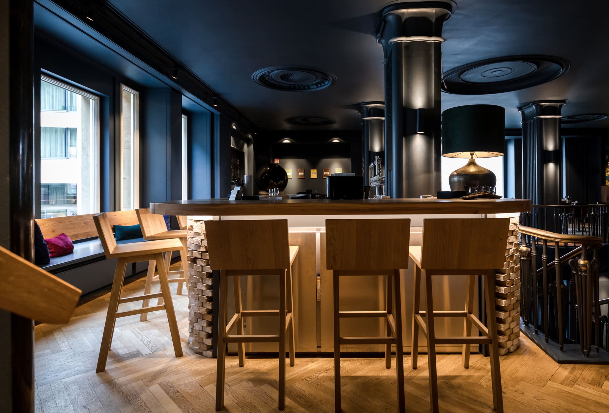 Bar counter with brick base and timber top, three pale bar stools, window bench with cushions at left, herringbone parquet