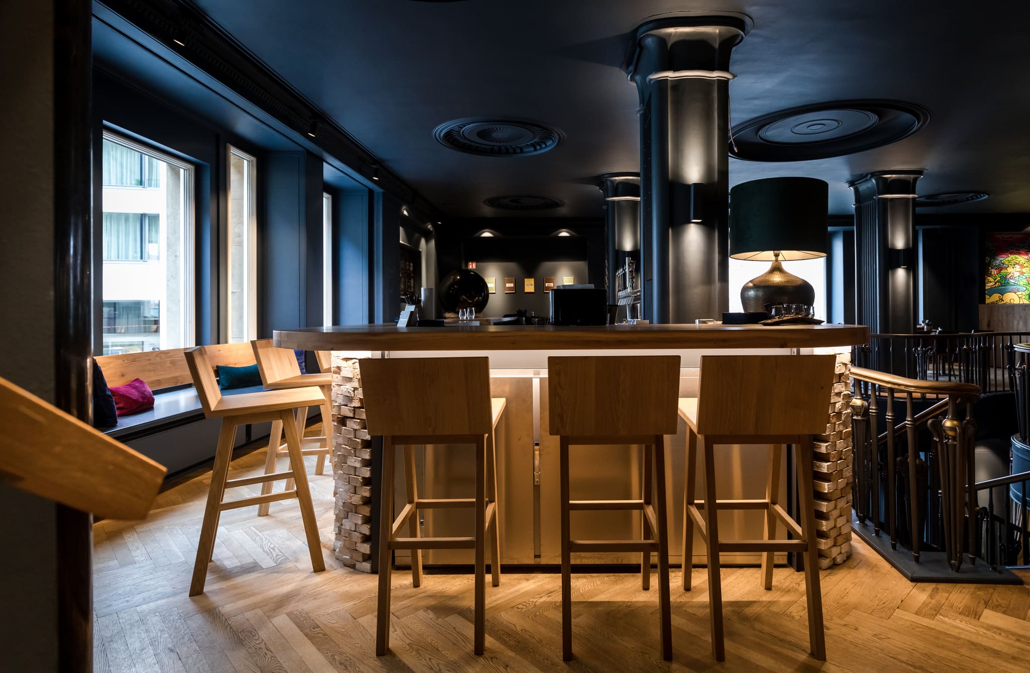 Bar counter with brick base and timber top, three pale bar stools, window bench with cushions at left, herringbone parquet