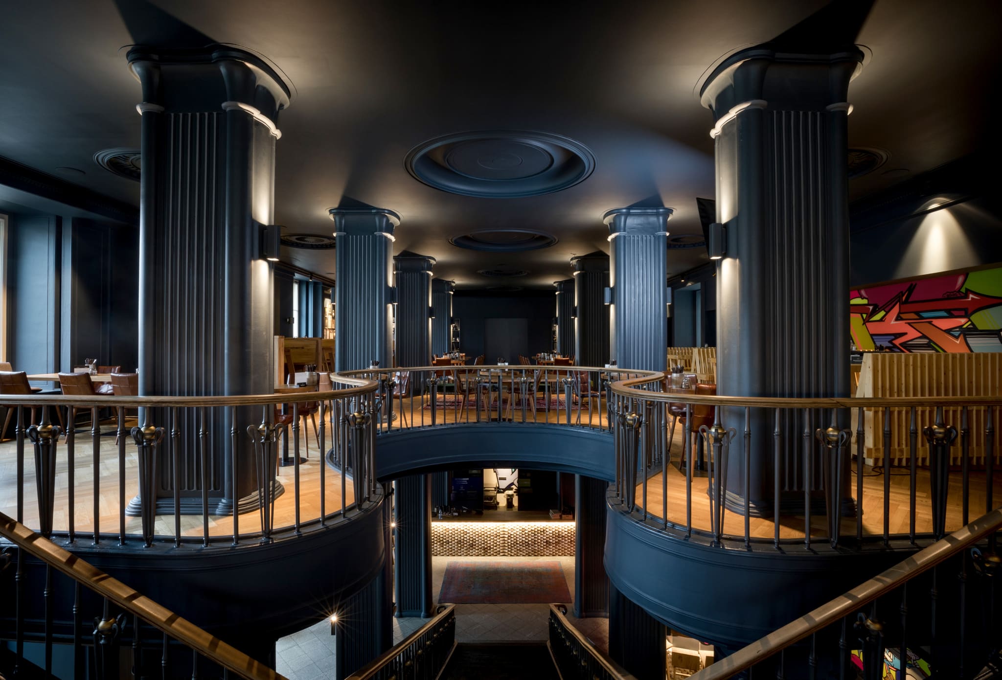 View from the stair head across the dining room, dark blue columns, gallery balustrade, tables with leather chairs, graffiti painting