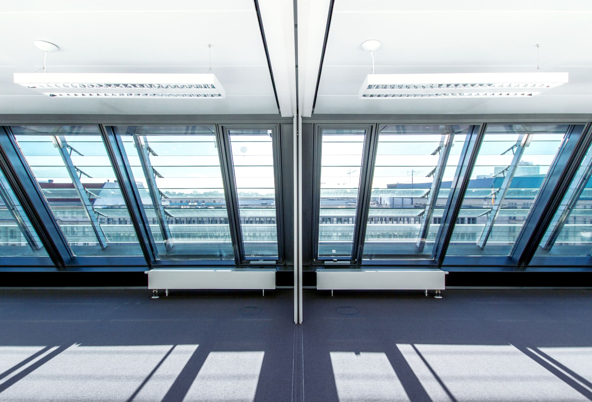 Empty room beneath inclined glazing with a view over rooftops, dark blue carpet, suspended luminaires, radiators below the windows