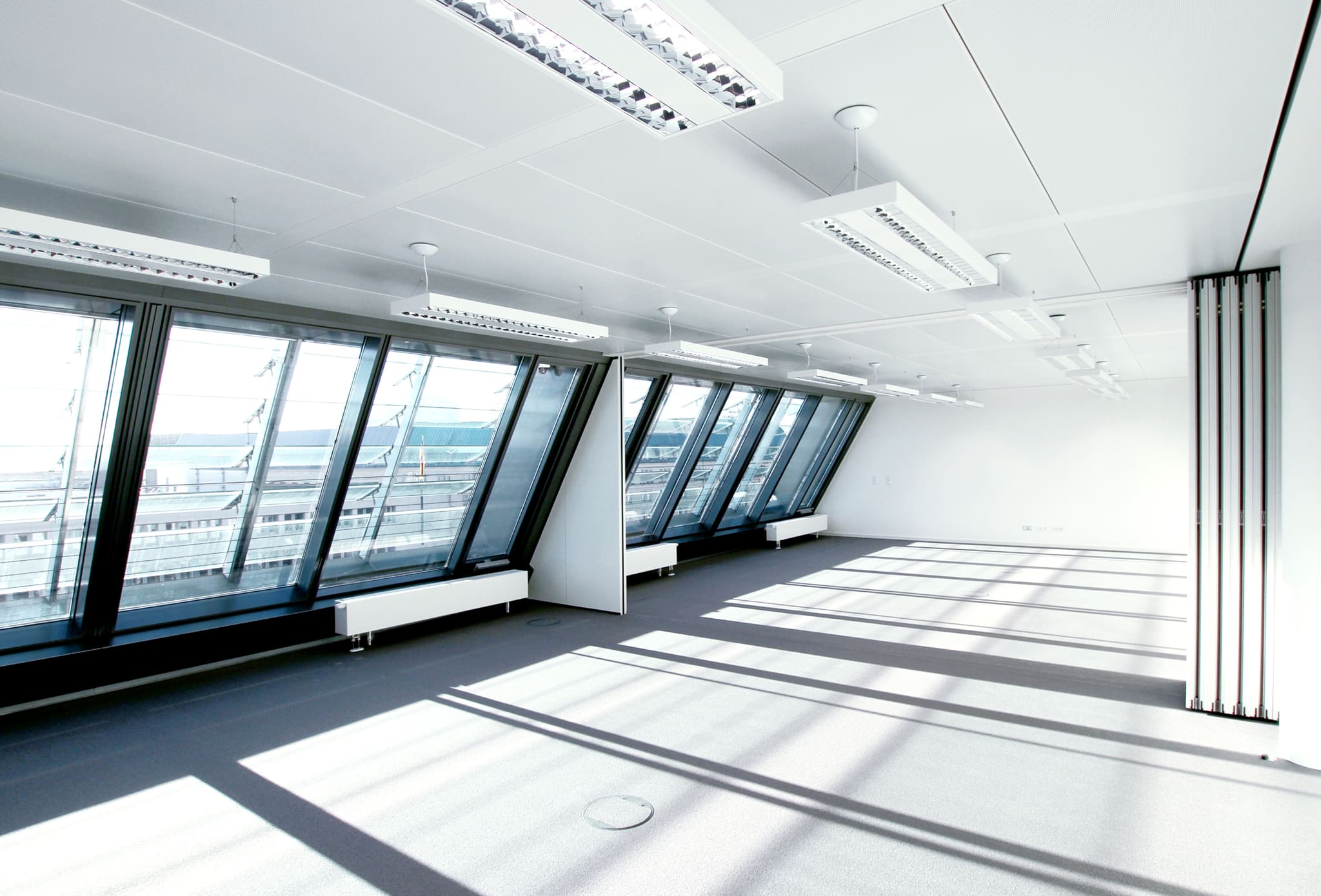 Empty bright hall, inclined glazing on the left, dark grey carpet, ceiling luminaires, stacked mobile partition wall on the right