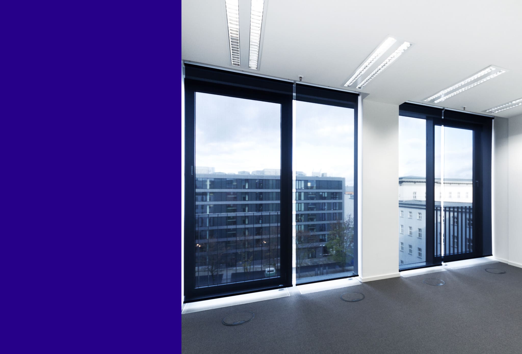 Empty office with floor-to-ceiling windows and lowered screen blinds, grey carpet with round floor box covers, ceiling luminaires