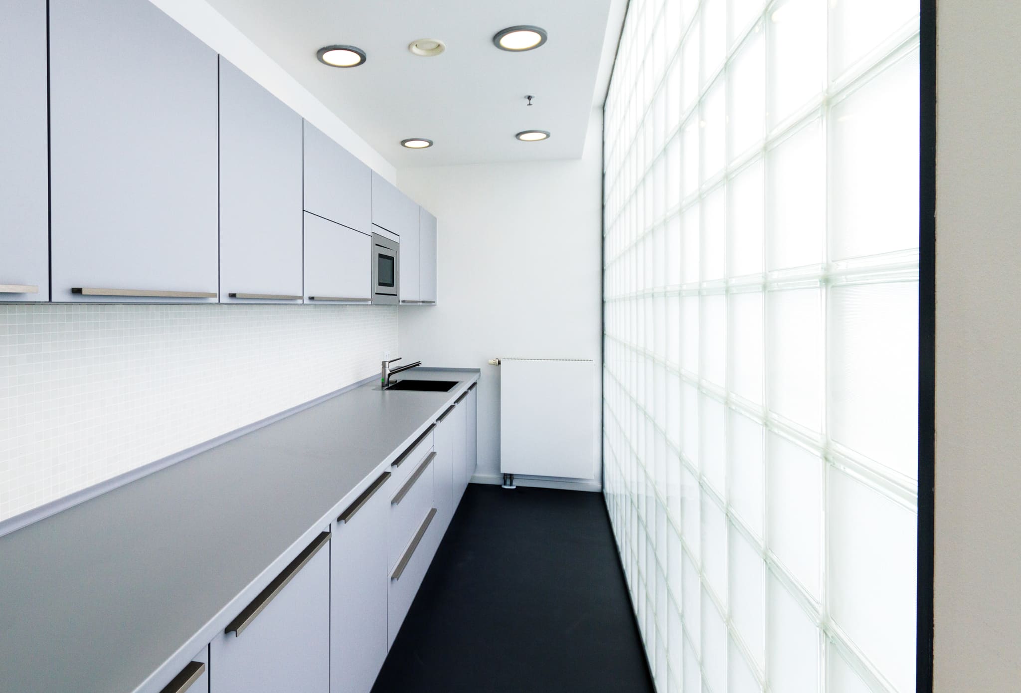 Narrow kitchenette with pale grey fronts, grey worktop, sink, mosaic tile splashback, dark floor and a glass block wall right