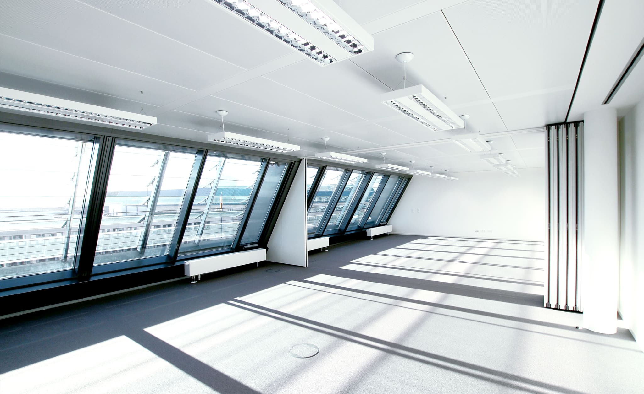 Empty open-plan space with a sloping glass façade, a white grid ceiling and stripes of light from the window profiles on the carpet