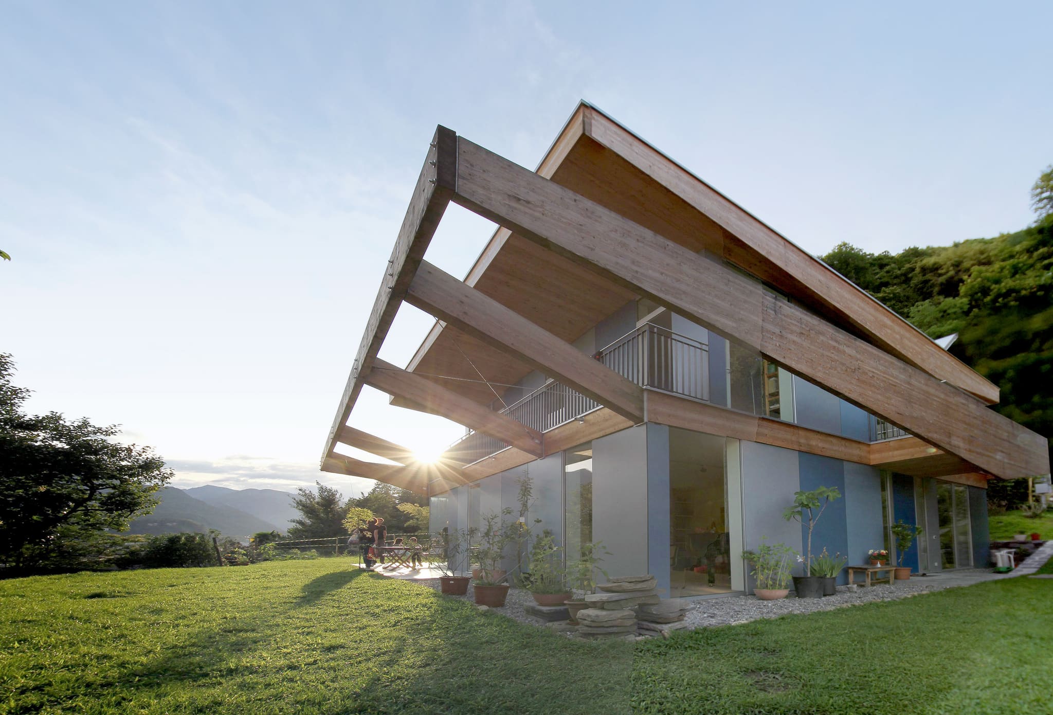 Two-storey house seen from the lawn, deeply cantilevered timber frame above the terrace, low sun, mountains behind