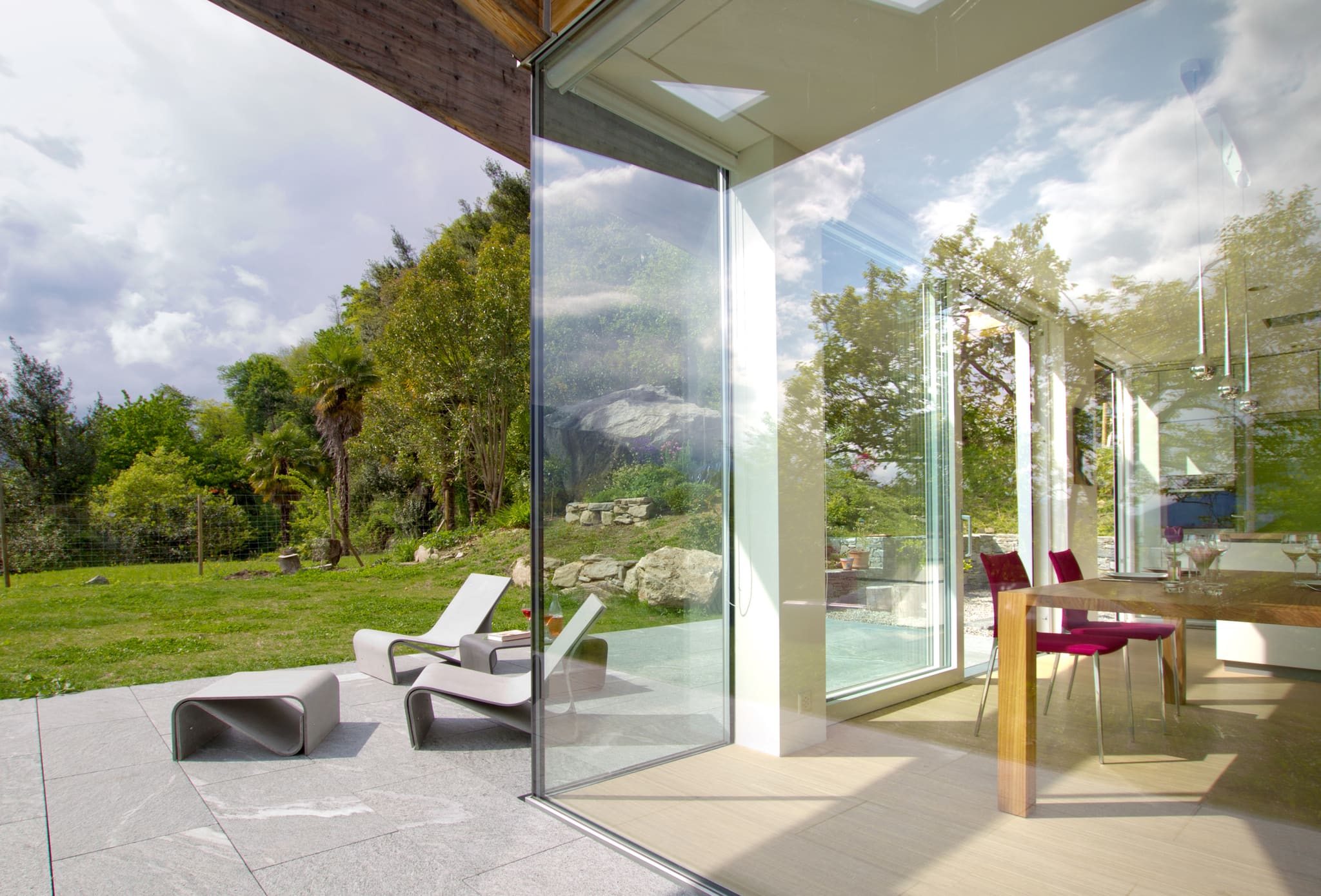 View from the stone terrace through frameless corner glazing into the dining area, loungers at left, garden and trees