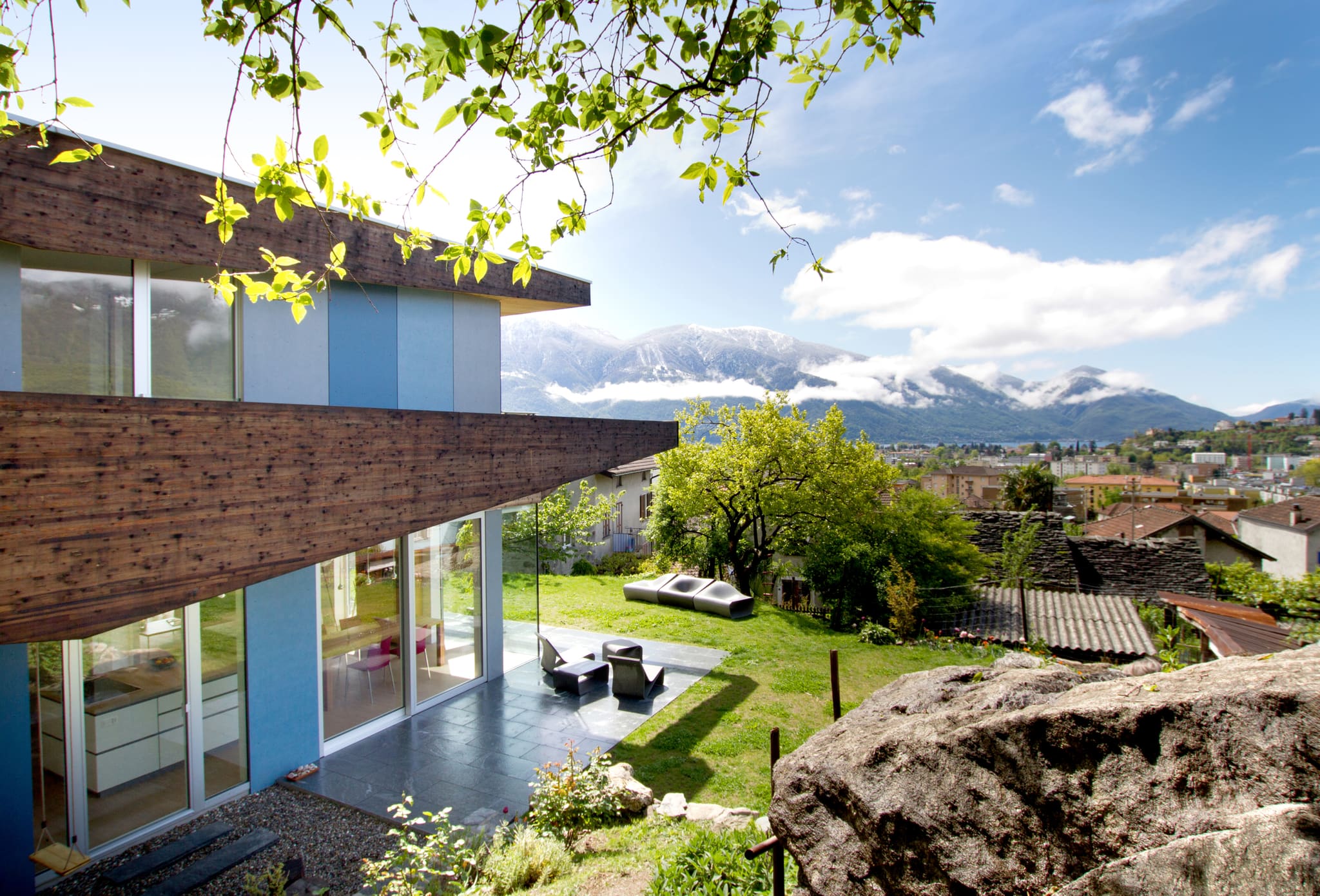 Corner of the house with blue façade panels and cantilevered timber frame, lawn with loungers, town, lake and mountains behind