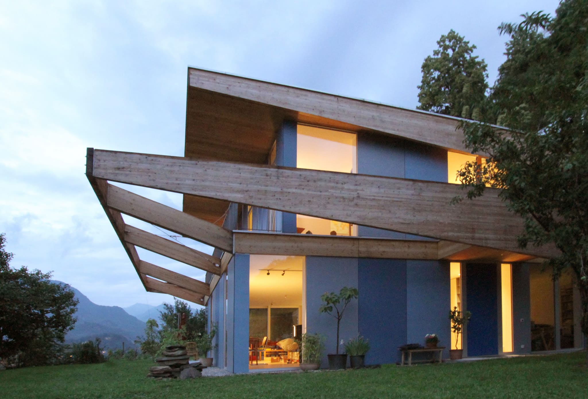 House at dusk, warmly lit interiors behind blue façade panels, tapering timber frame projecting above the terrace