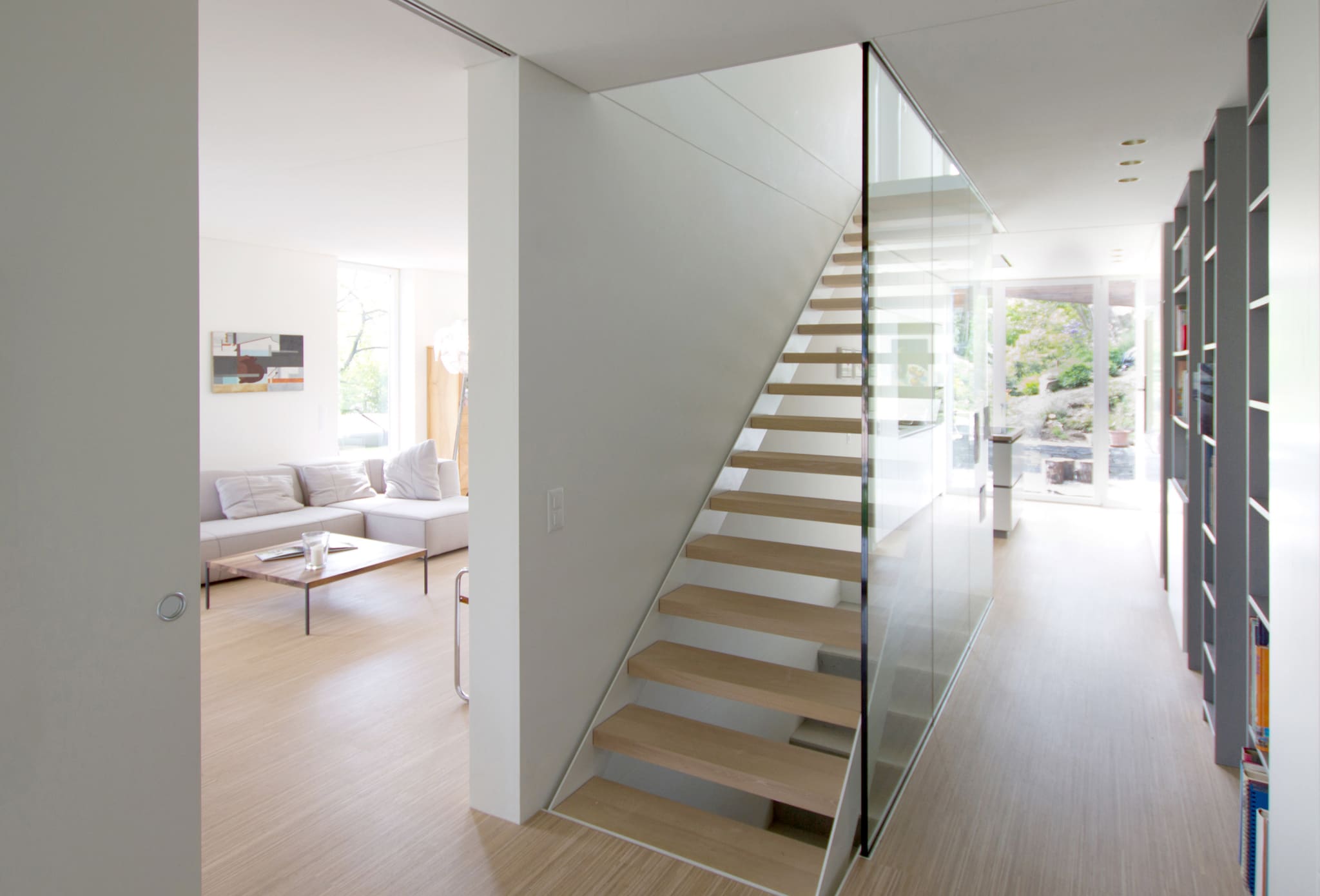 Interior with open timber stair behind a full-height glass balustrade, living area at left, bookshelves at right, garden beyond