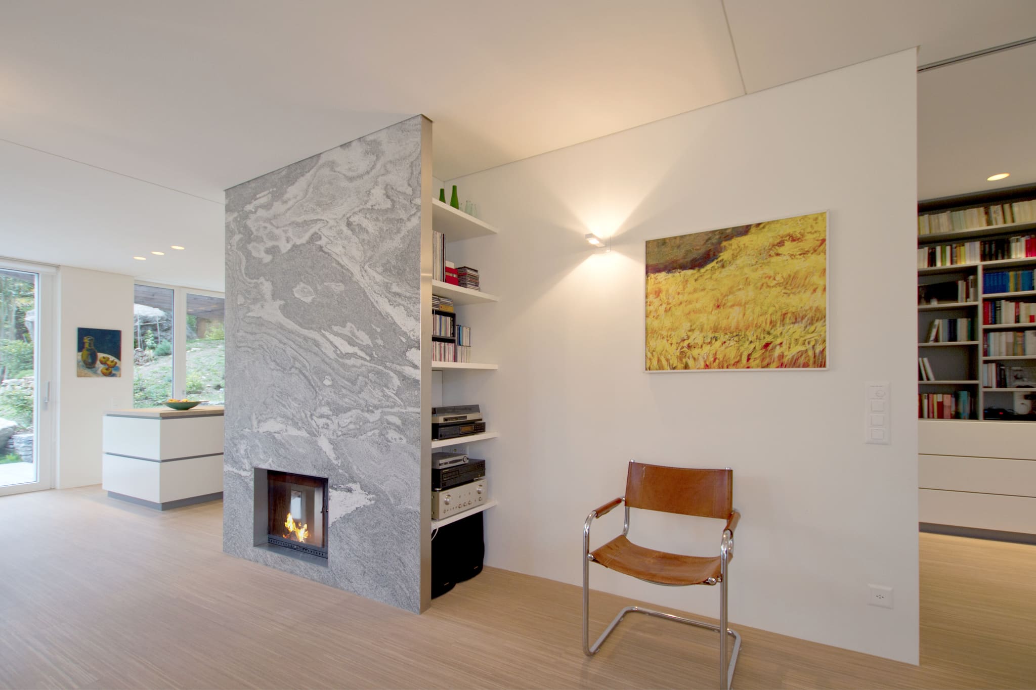 Freistehender Kaminblock aus grauem Naturstein mit seitlichen Regalfächern, rechts Gemälde und Bücherwand, links offene Küche am Fenster