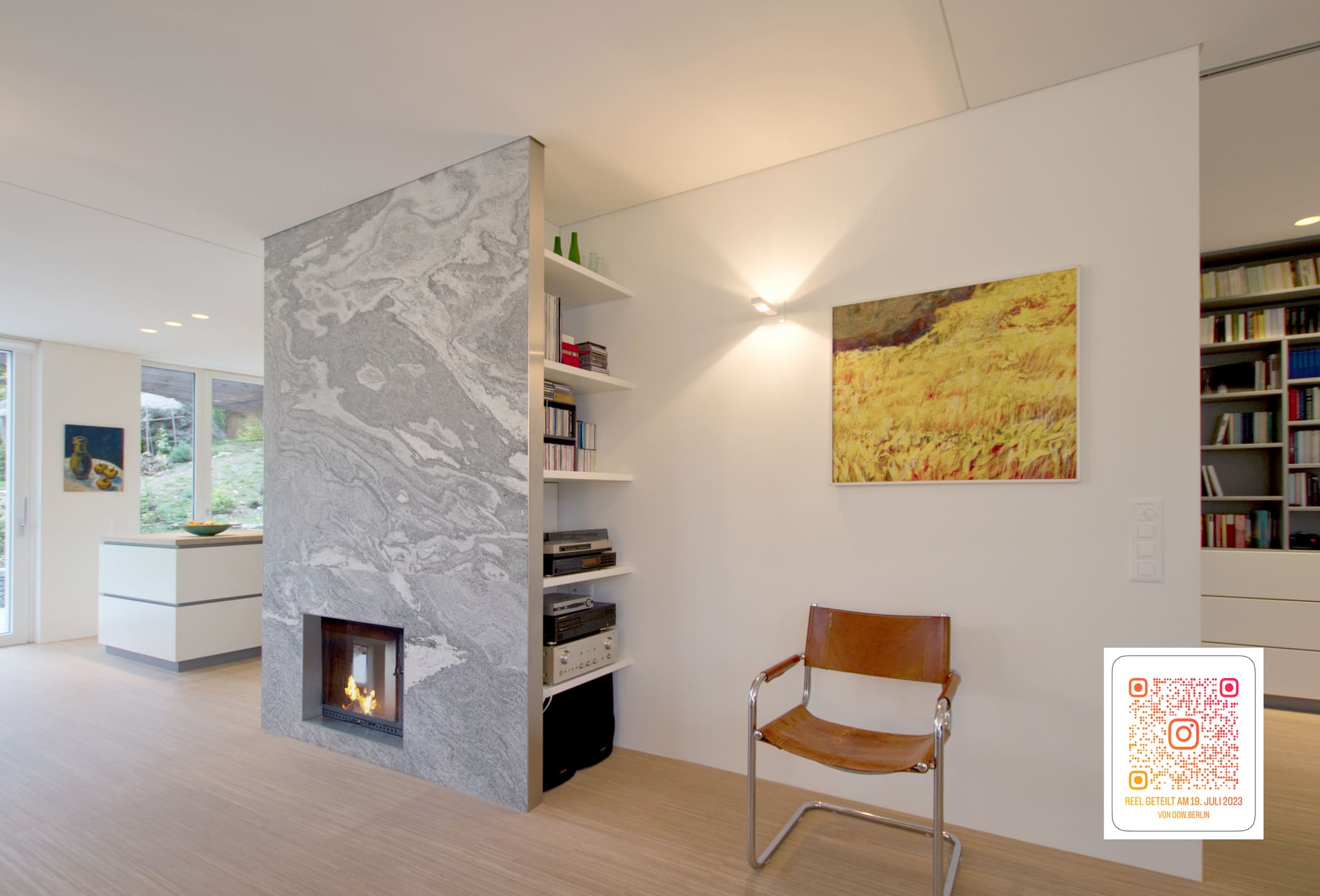 Free-standing fireplace block of grey natural stone with side shelving, painting and book wall at right, open kitchen at left