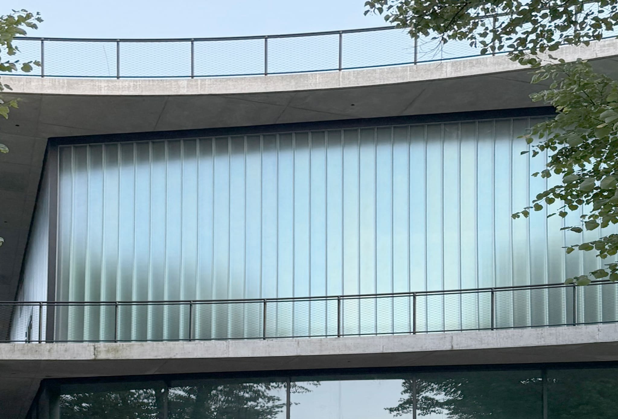 Fassadenausschnitt mit milchiger Profilglaswand zwischen zwei Betondecken, davor Stahlgeländer mit Drahtnetz, rechts Baumzweige