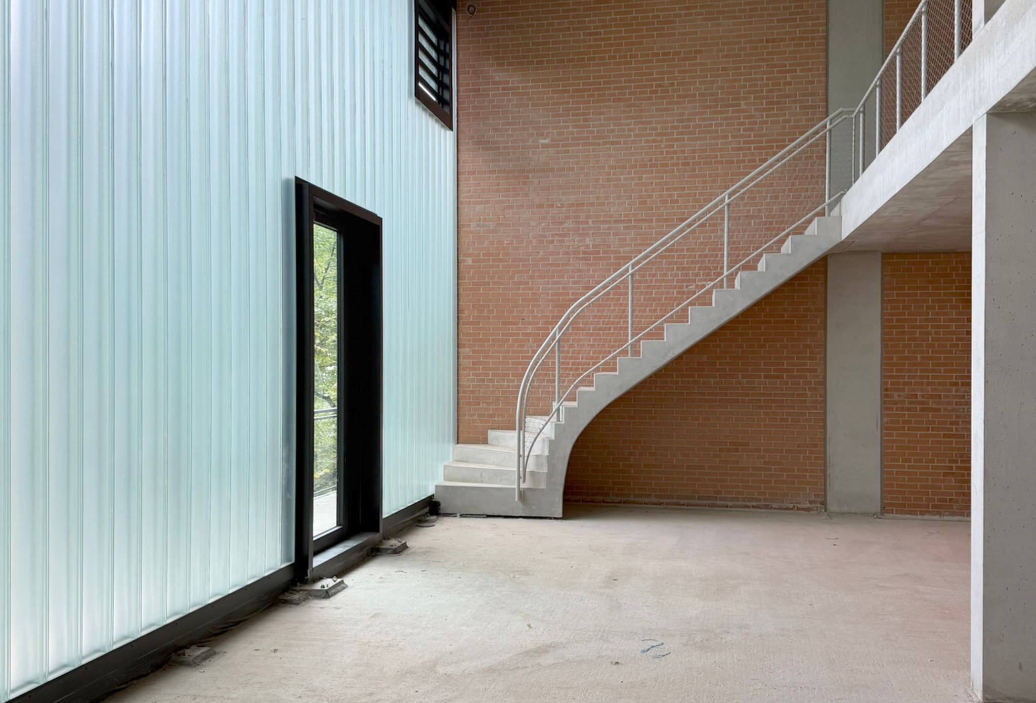 Rohbauraum mit milchiger Profilglaswand links, roter Ziegelwand rechts und freitragender Betontreppe mit schlankem Rohrhandlauf zur Galerie