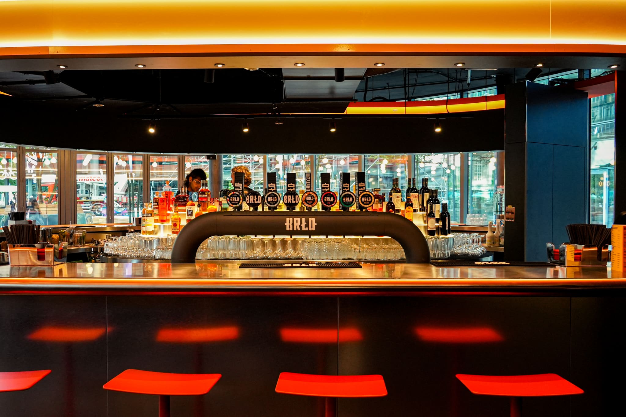 Stainless steel bar with beer taps and bottle shelf, red stools in front, glazed facade behind, warm light band above