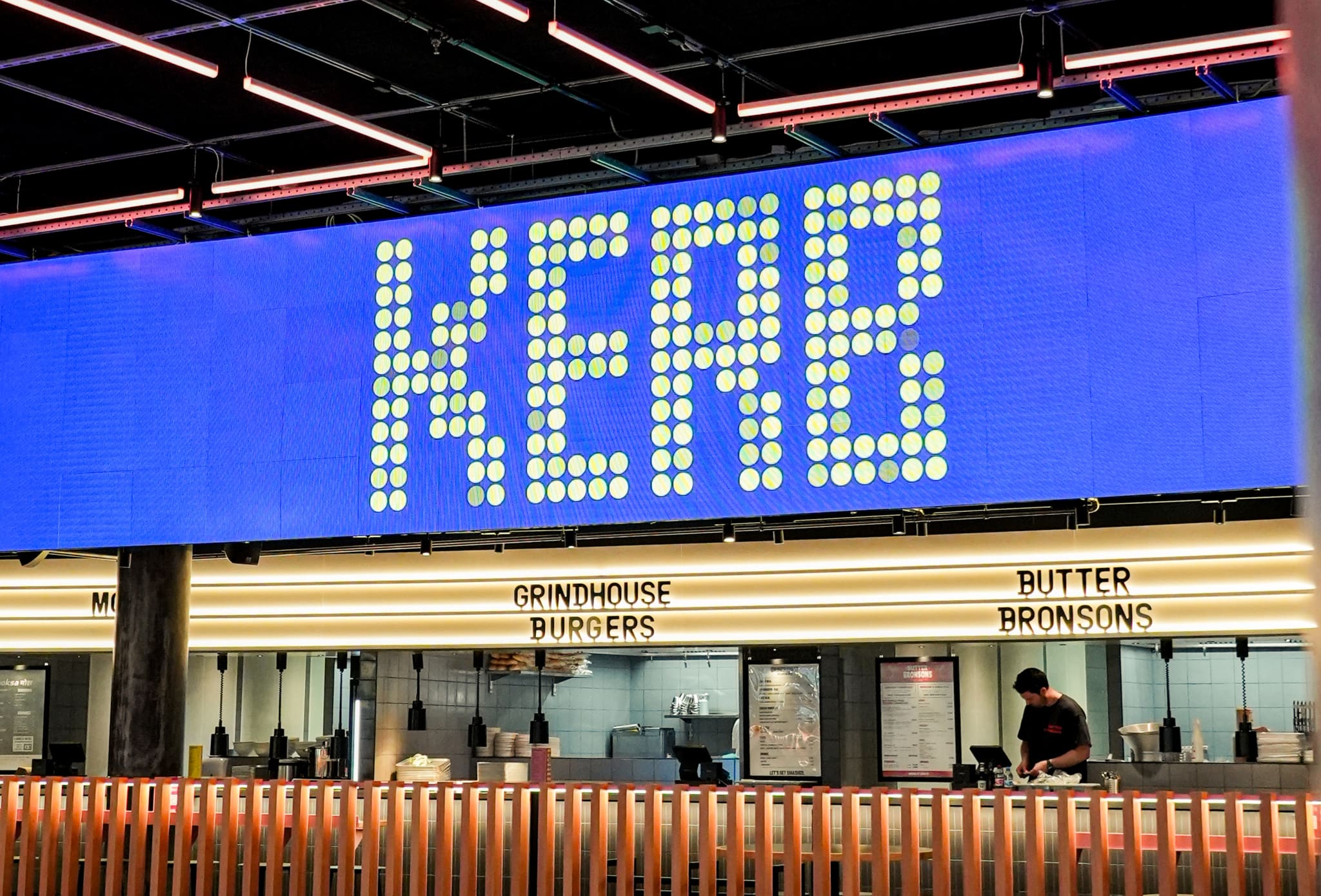 Großes blaues LED-Display über einer Reihe von Foodständen mit beleuchteten Schilderbändern, davor eine Brüstung aus Holzlamellen